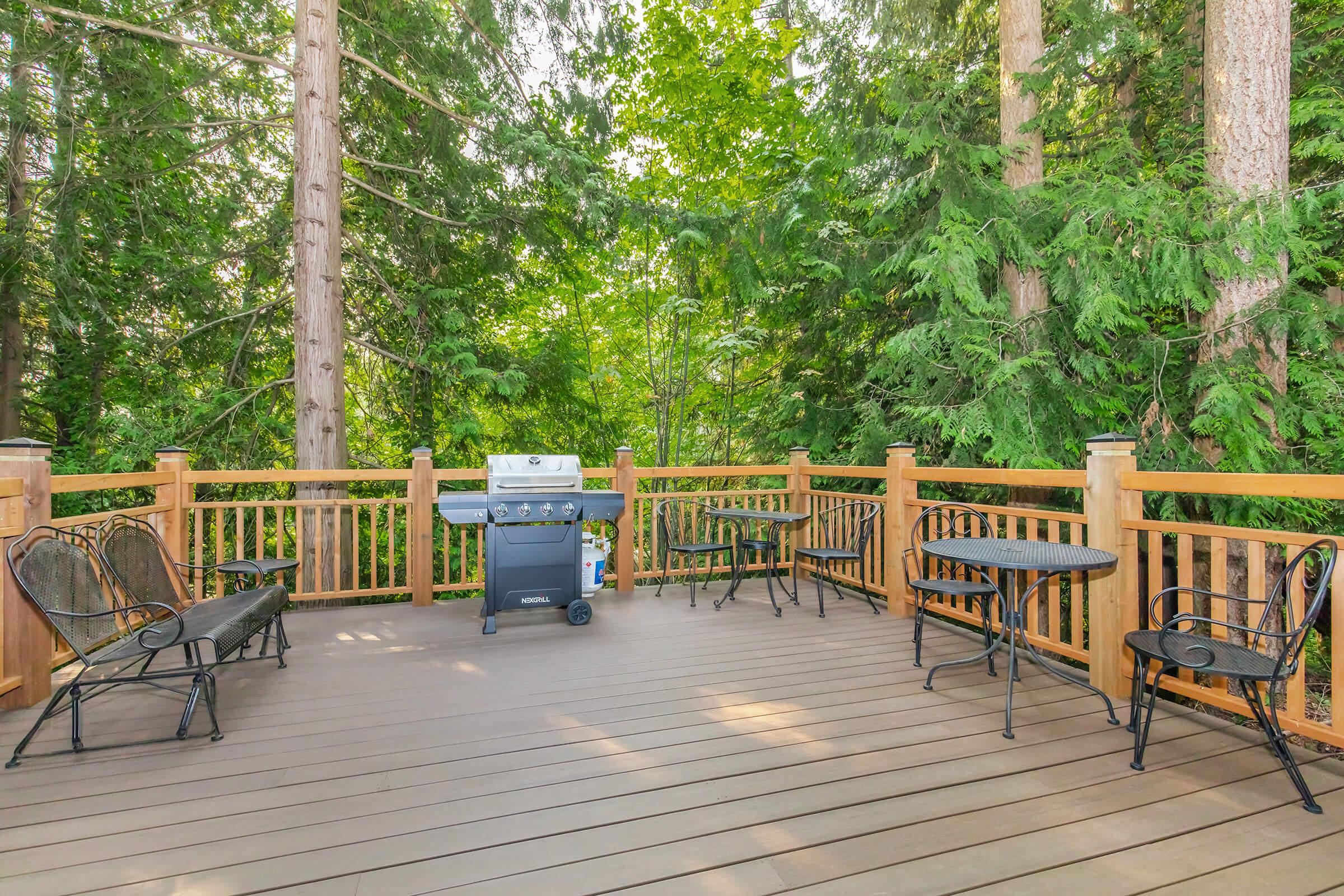 A spacious outdoor deck surrounded by tall trees, featuring a barbecue grill, several black metal chairs, and small tables. The wooden railing adds a warm touch to the natural setting, creating an inviting space for gatherings or relaxing in a serene, green environment.