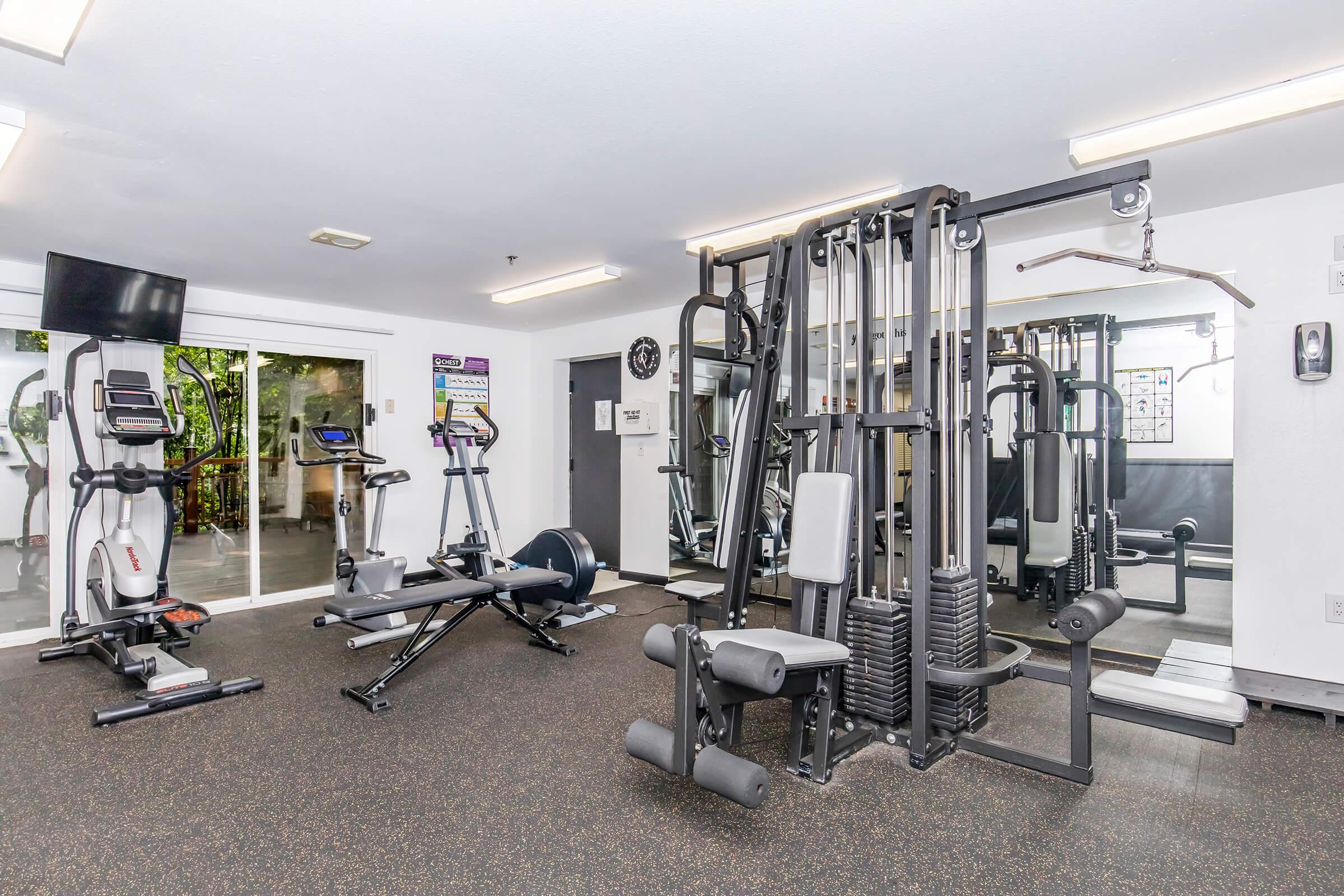 A well-equipped gym interior featuring various workout machines, including a cable machine, elliptical, and stationary bike. Large mirrors reflect the space, and there's a television mounted on the wall. The area is well-lit with overhead lighting, providing a clean and modern atmosphere for exercise.