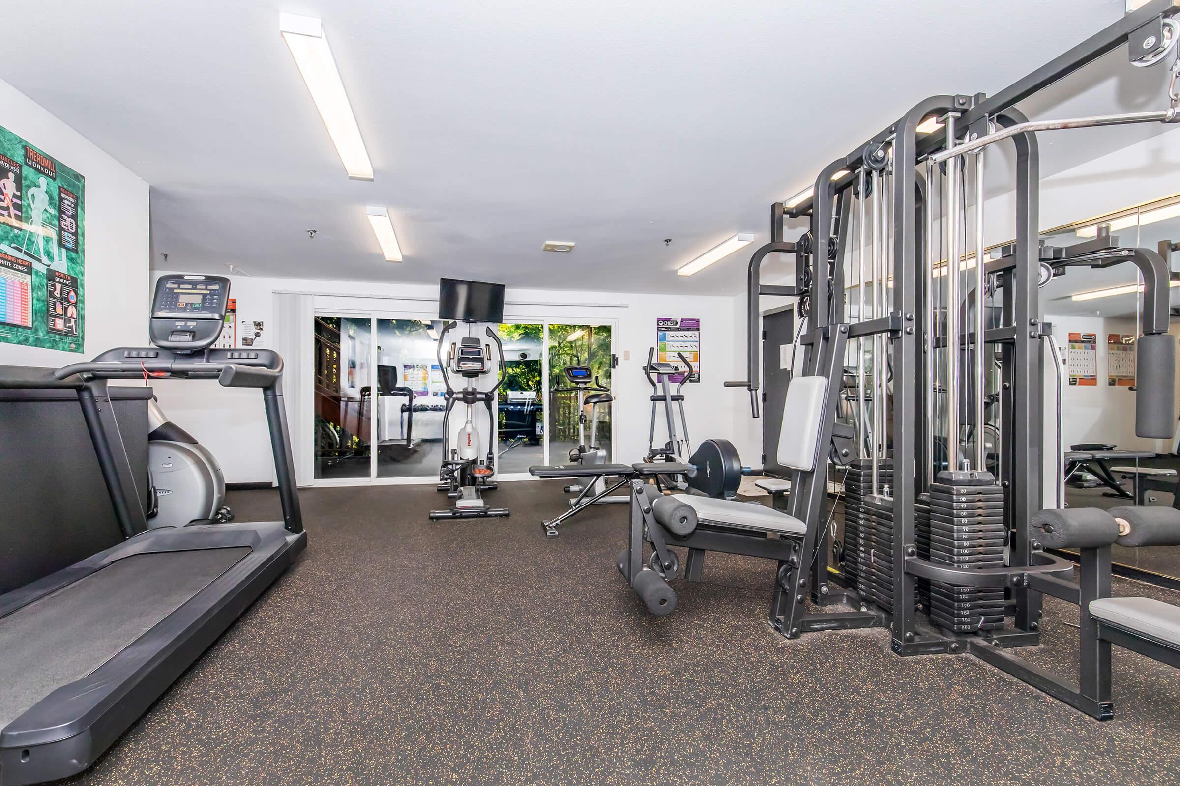 A well-equipped gym featuring a treadmill, an elliptical machine, and a multi-station weight machine. The area is brightly lit with overhead fluorescent lights and has rubber flooring. Large windows offer a view to the outside, creating an inviting and spacious workout environment.