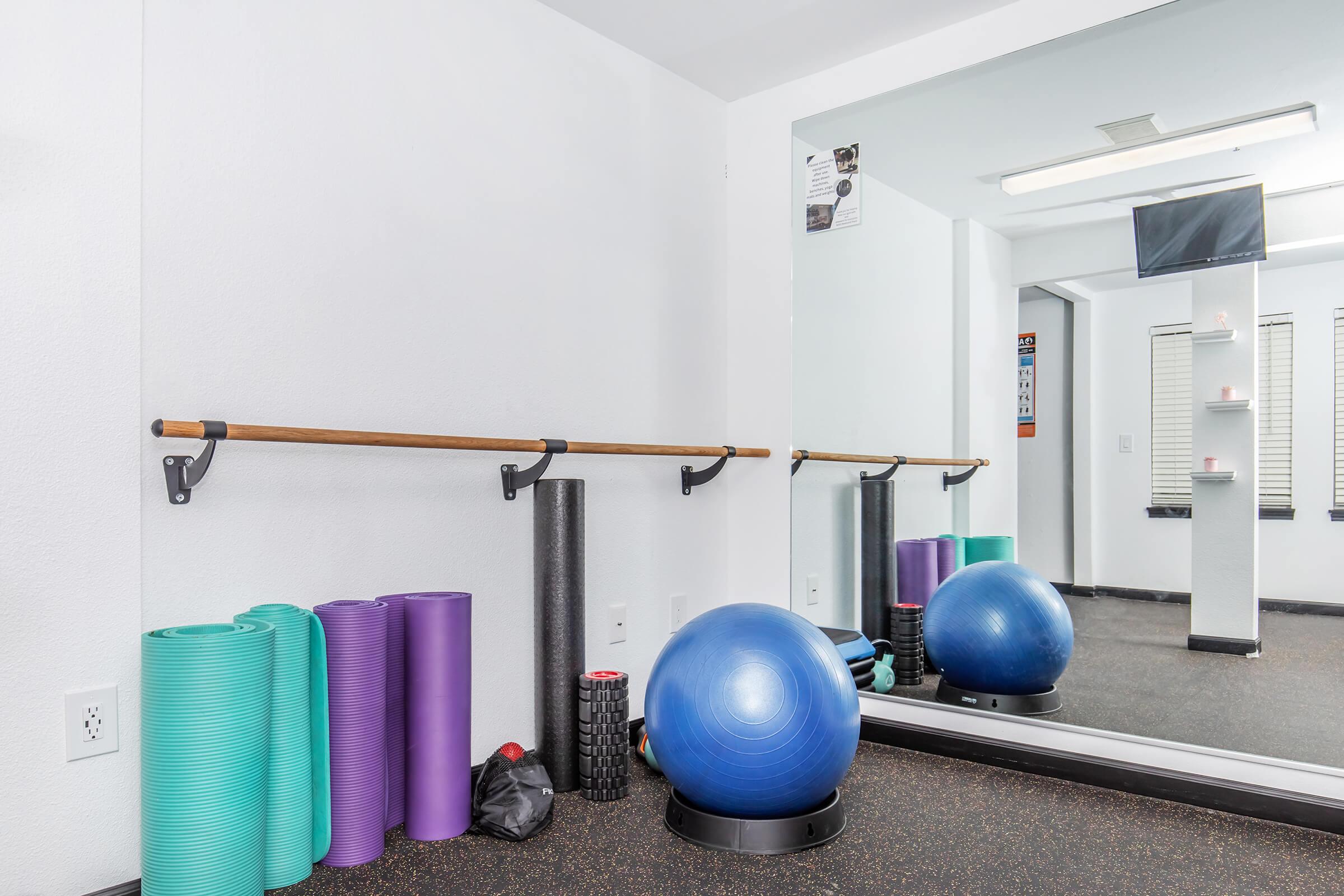 A fitness studio featuring exercise equipment. There are colorful rolld-up yoga mats in purple and teal, a blue stability ball, a black foam roller, and a wooden barre against a mirrored wall. The room has a television and candles on a shelf, creating a tranquil workout environment.