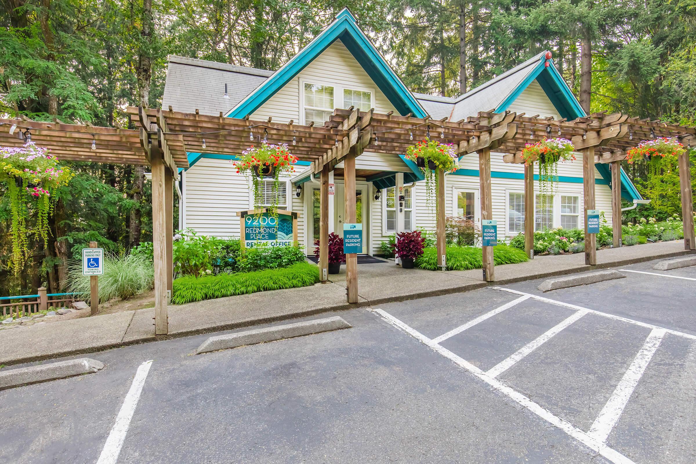 A charming building surrounded by greenery, featuring a white façade with teal trim. It has a wooden trellis adorned with hanging flower baskets in vibrant colors. The entrance is inviting, with signage indicating it might be a welcoming space like an office or retreat. The asphalt parking area includes marked spaces.