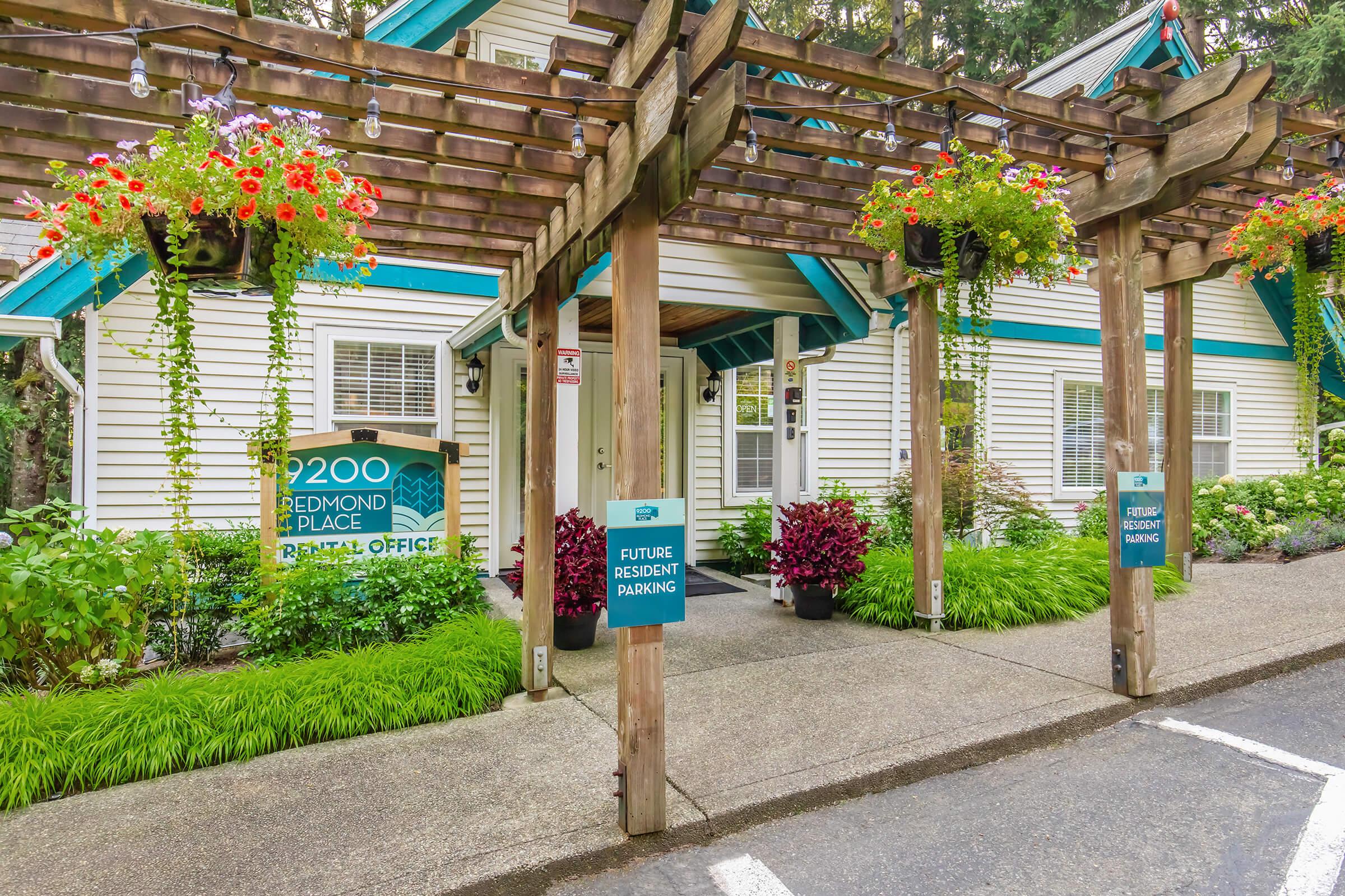 A view of the Redmond Place rental office entrance. The building features a wooden pergola adorned with colorful hanging flower baskets. Signs indicate the office number (2200) and future resident parking. The surrounding area is landscaped with vibrant plants and greenery.