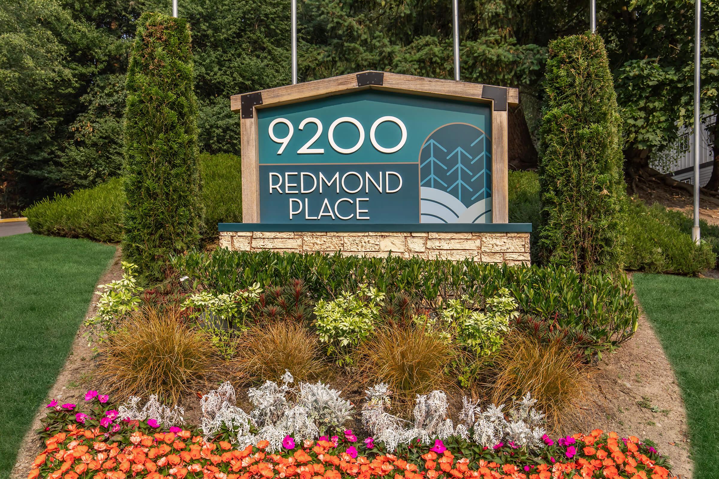 Sign displaying "9200 Redmond Place" surrounded by landscaped greenery and colorful flower beds. Flags are visible in the background, indicating the entrance to a property or location.