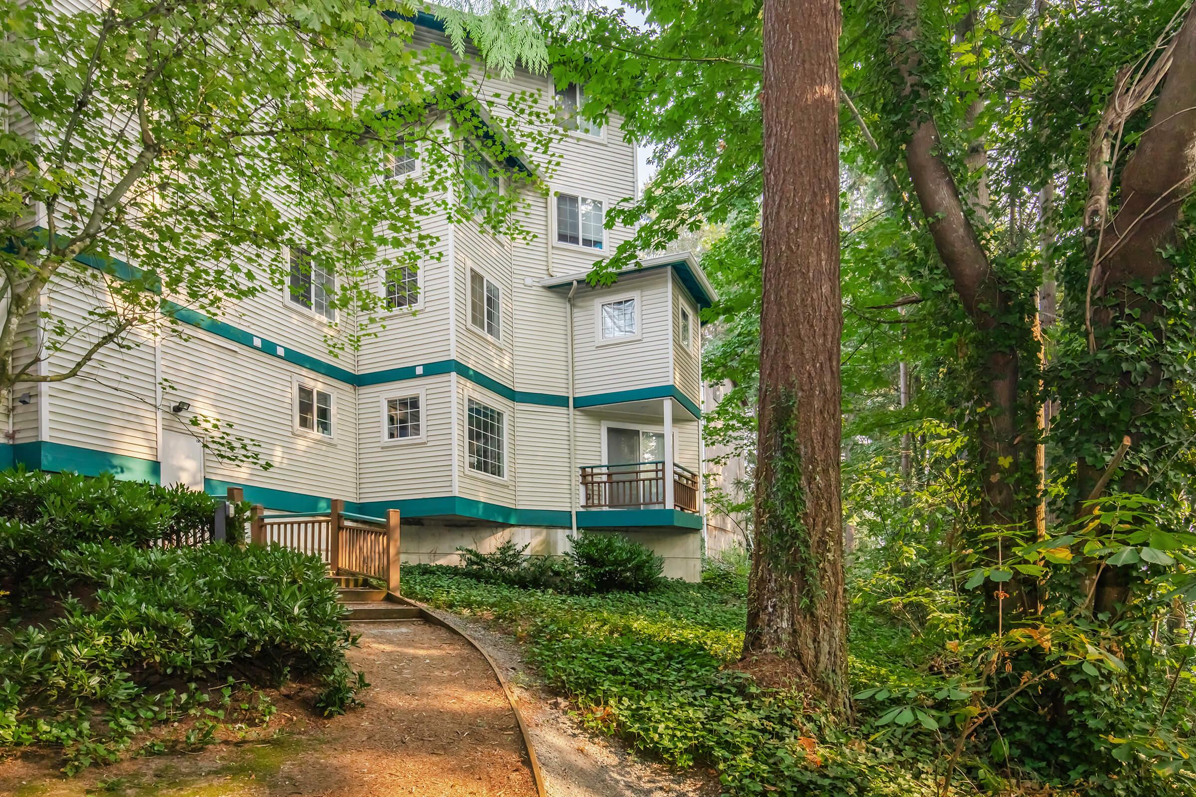A corner view of a multi-story house surrounded by lush greenery. The exterior features light-colored siding with teal accents, set amidst tall trees and shrubbery. A winding path leads from the foreground to the house, inviting visitors to explore the serene, wooded environment.