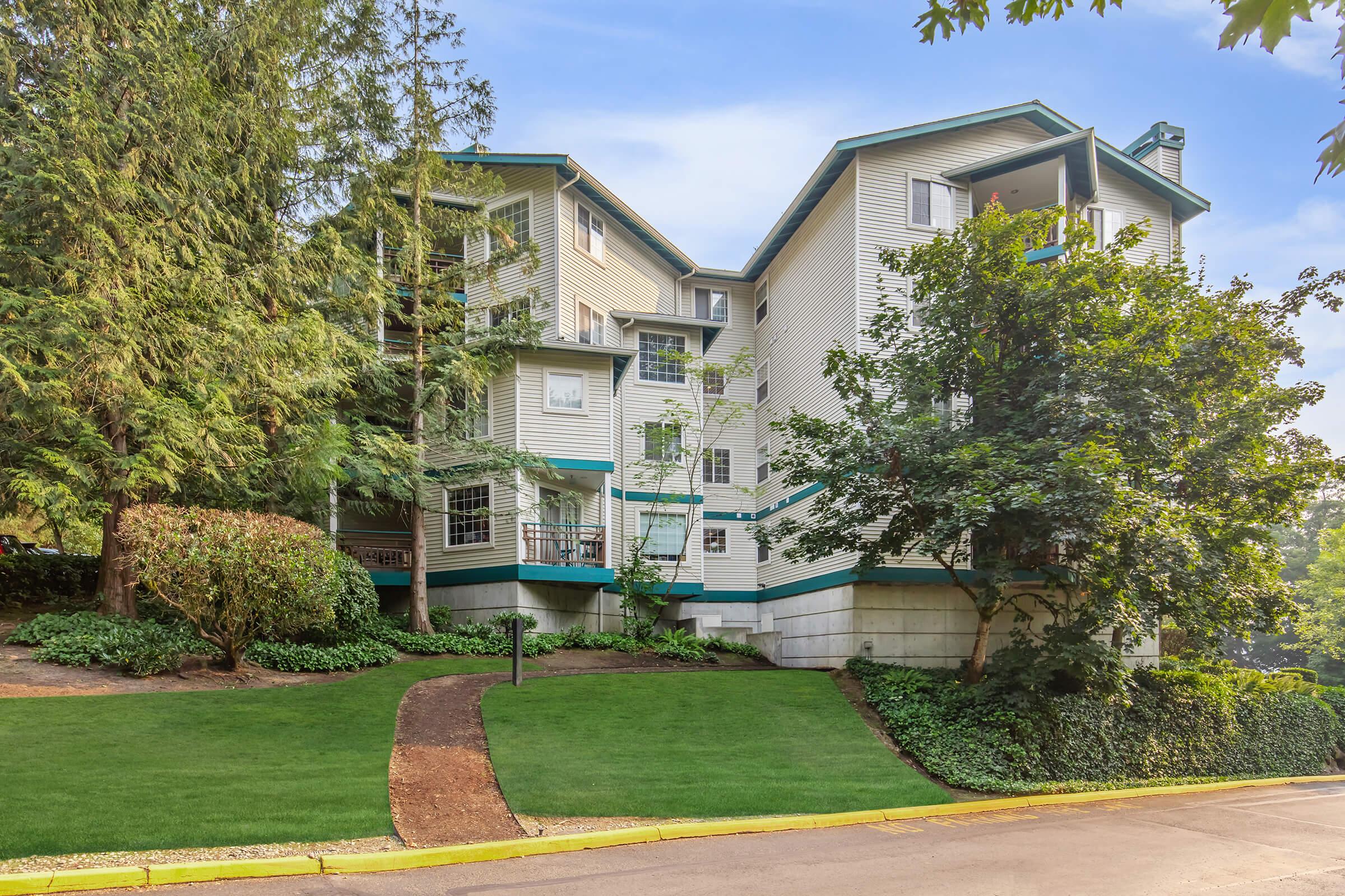 A multi-story residential building surrounded by lush greenery and well-maintained landscaping. The structure features a light-colored exterior with teal accents, and there are trees and bushes enhancing the natural setting. A paved path leads up to the building entrance.