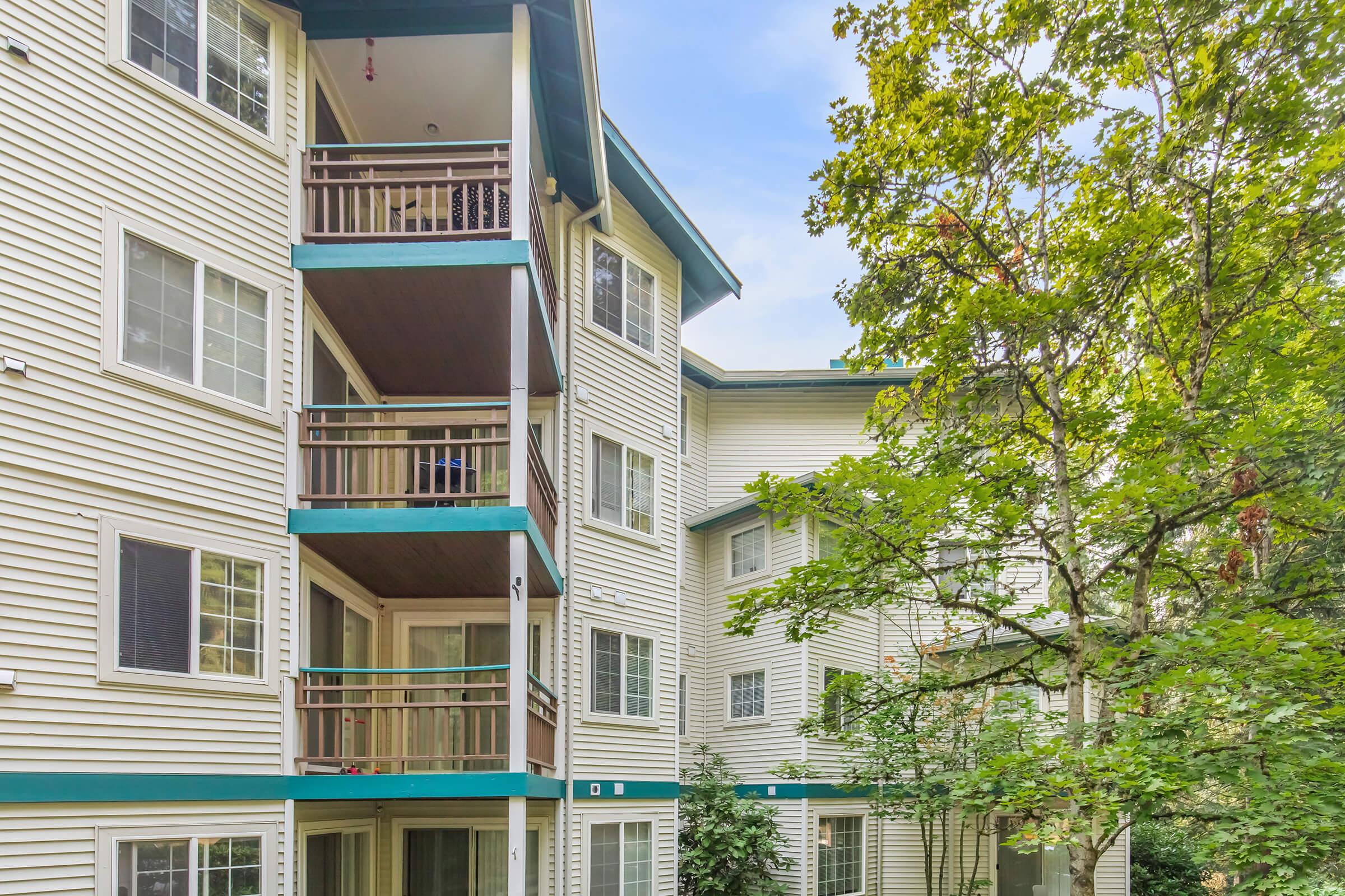 A multi-story apartment building with light-colored siding and green trim. Balconies are visible on multiple levels, surrounded by trees and shrubbery. The sky is clear, creating a bright atmosphere.