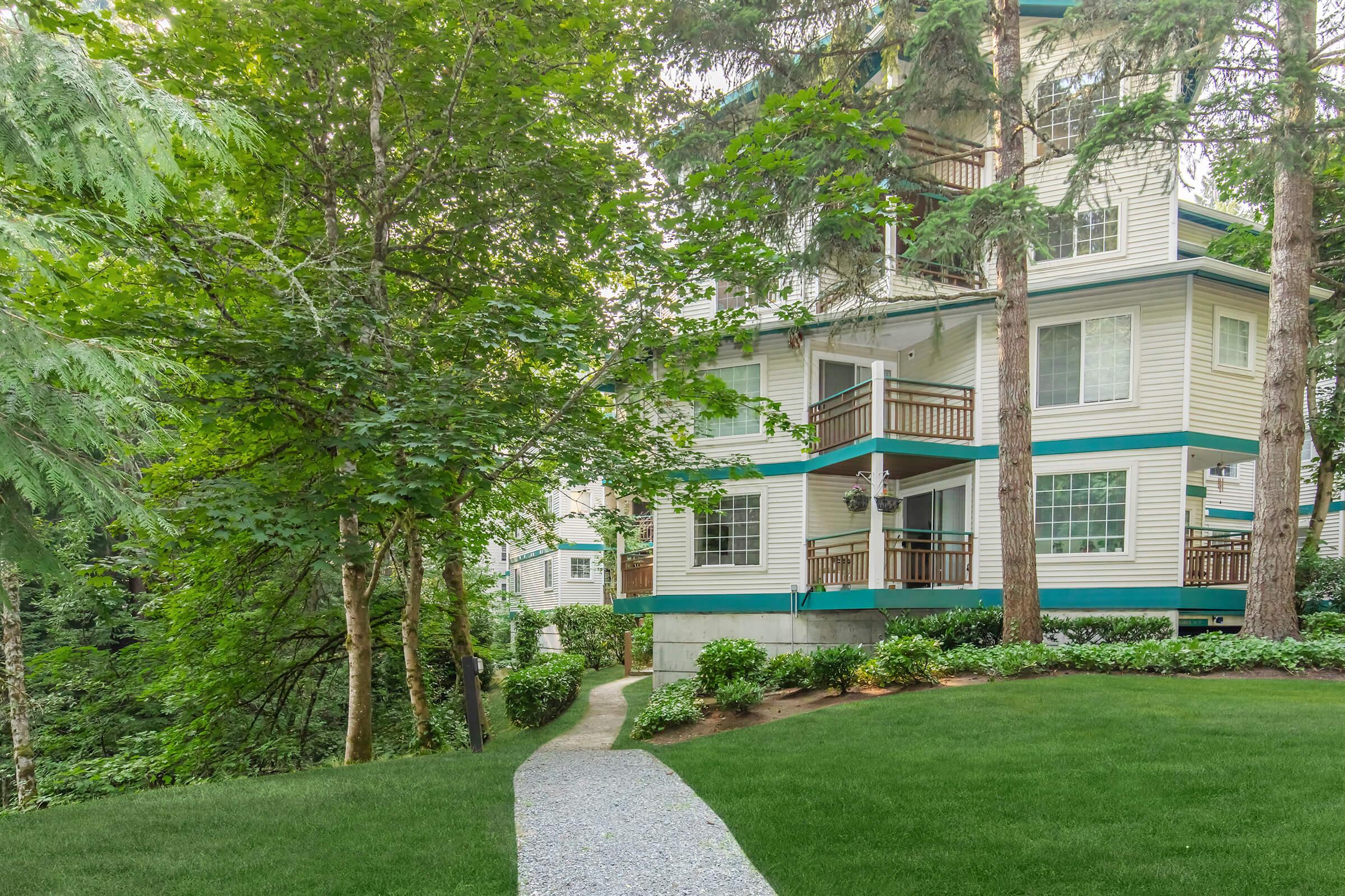 A landscaped path leading to a multi-story residential building surrounded by lush greenery and tall trees. The building features balconies with wooden railings and large windows. Soft grass and well-maintained hedges complement the serene, natural setting.