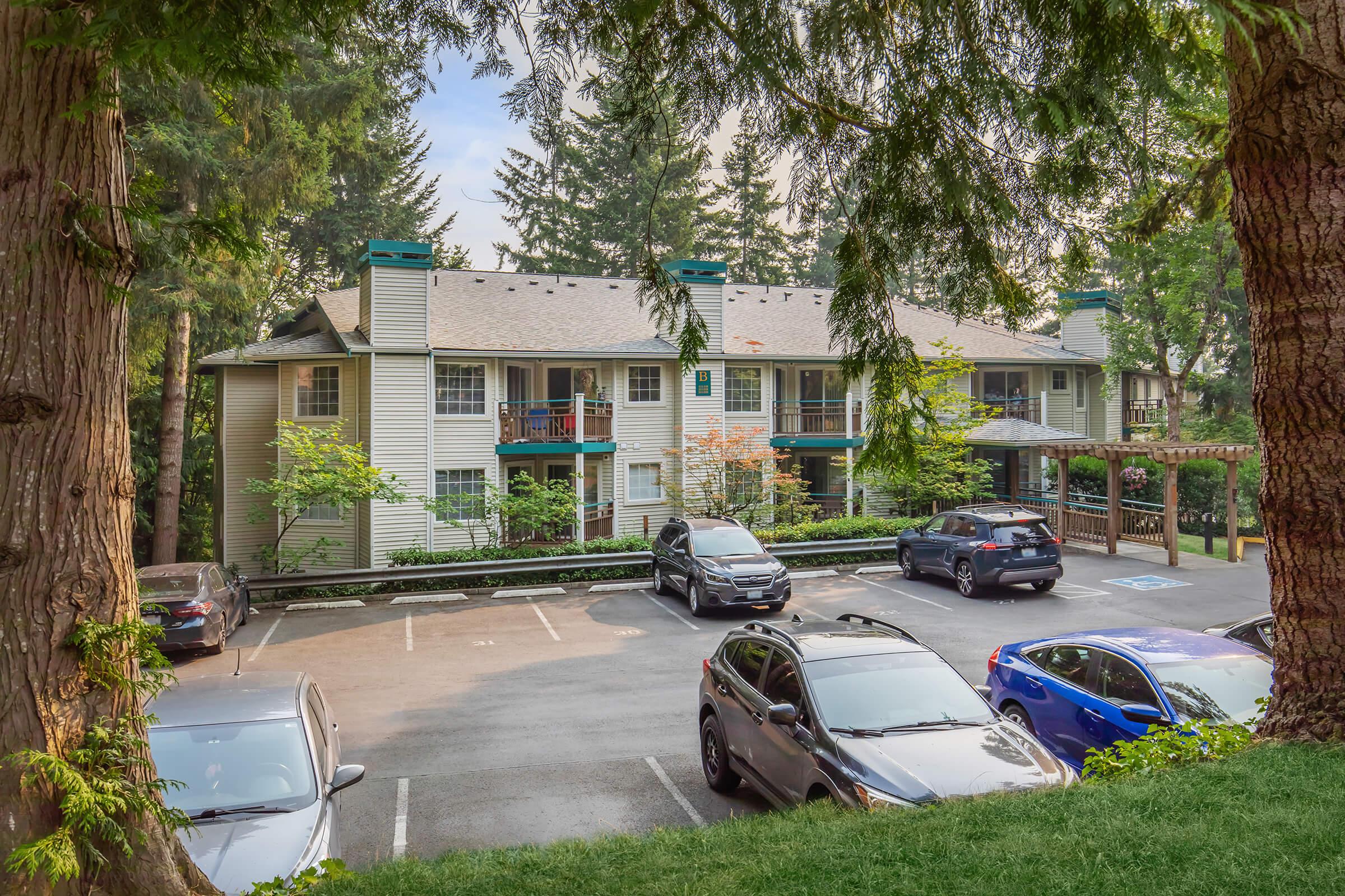 View of a residential apartment complex surrounded by trees. The building features multiple balconies and is set back from the parking area, which has several parked cars. The scene is sunny and green, suggesting a tranquil suburban environment.