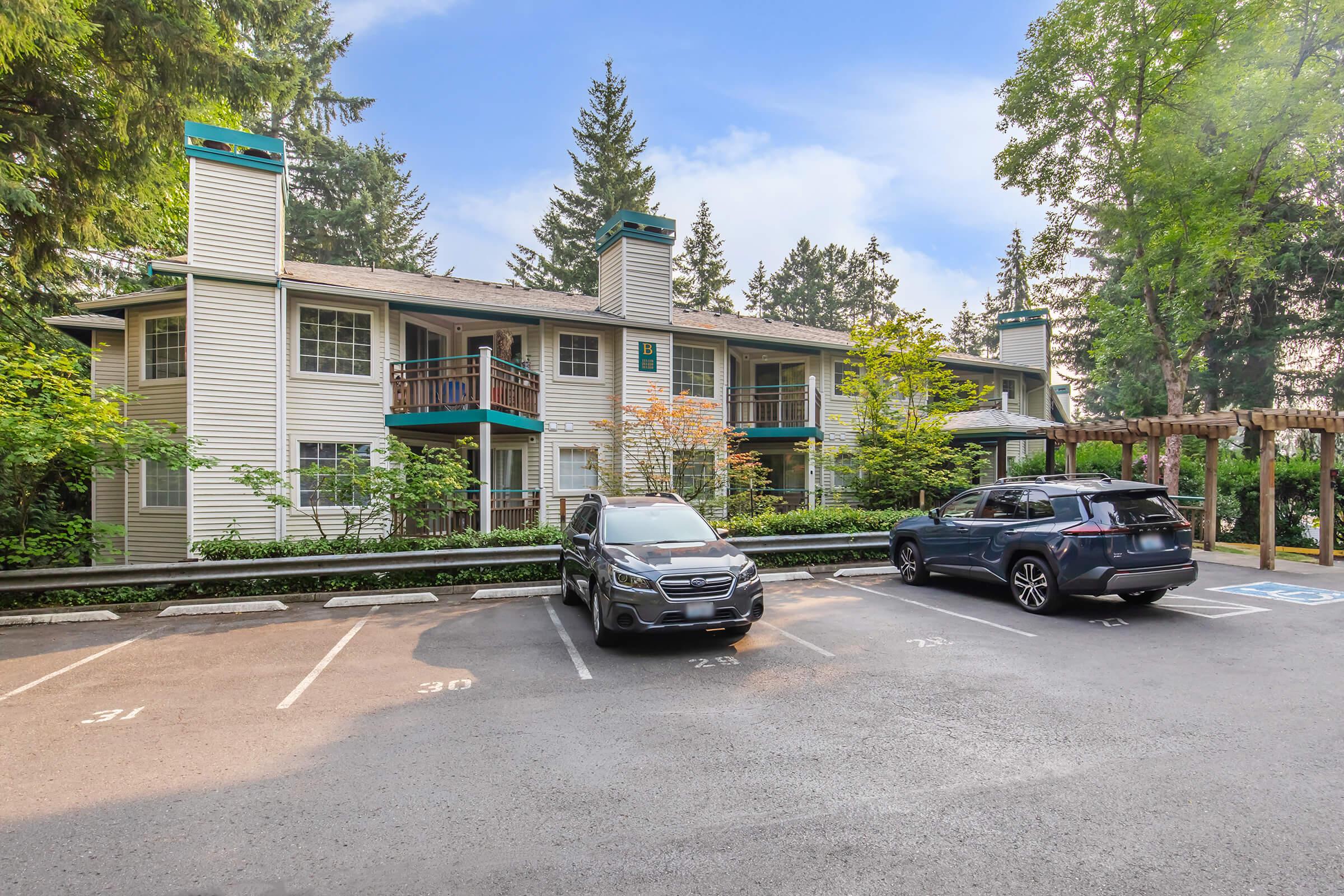 A residential apartment complex surrounded by greenery. The building features multiple balconies with a light-colored exterior and green accents. Two parked cars are visible in the foreground, alongside designated parking spaces. Tall trees provide a natural backdrop to the scene.
