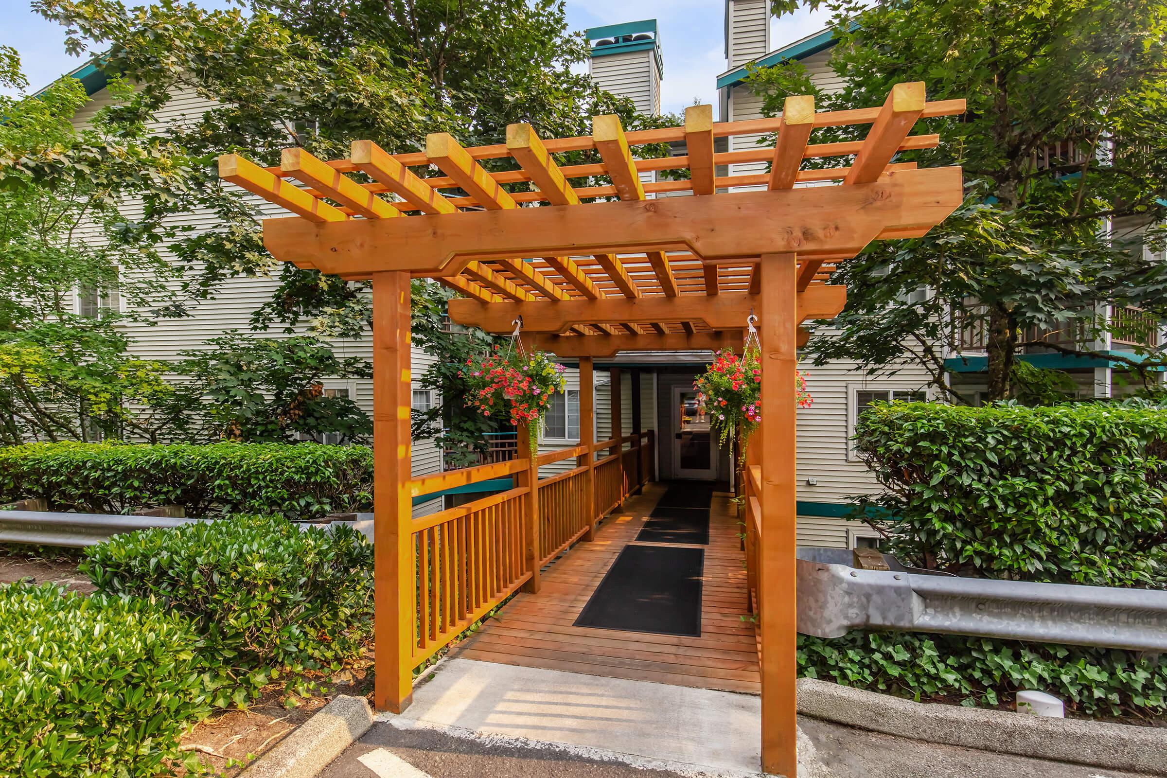 A wooden pergola covered with hanging plants frames a ramp leading to a building entrance. The ramp is surrounded by lush greenery, including neatly trimmed bushes, and the background features a multi-story building with light-colored siding. The scene is bright and inviting, suggesting a welcoming atmosphere.