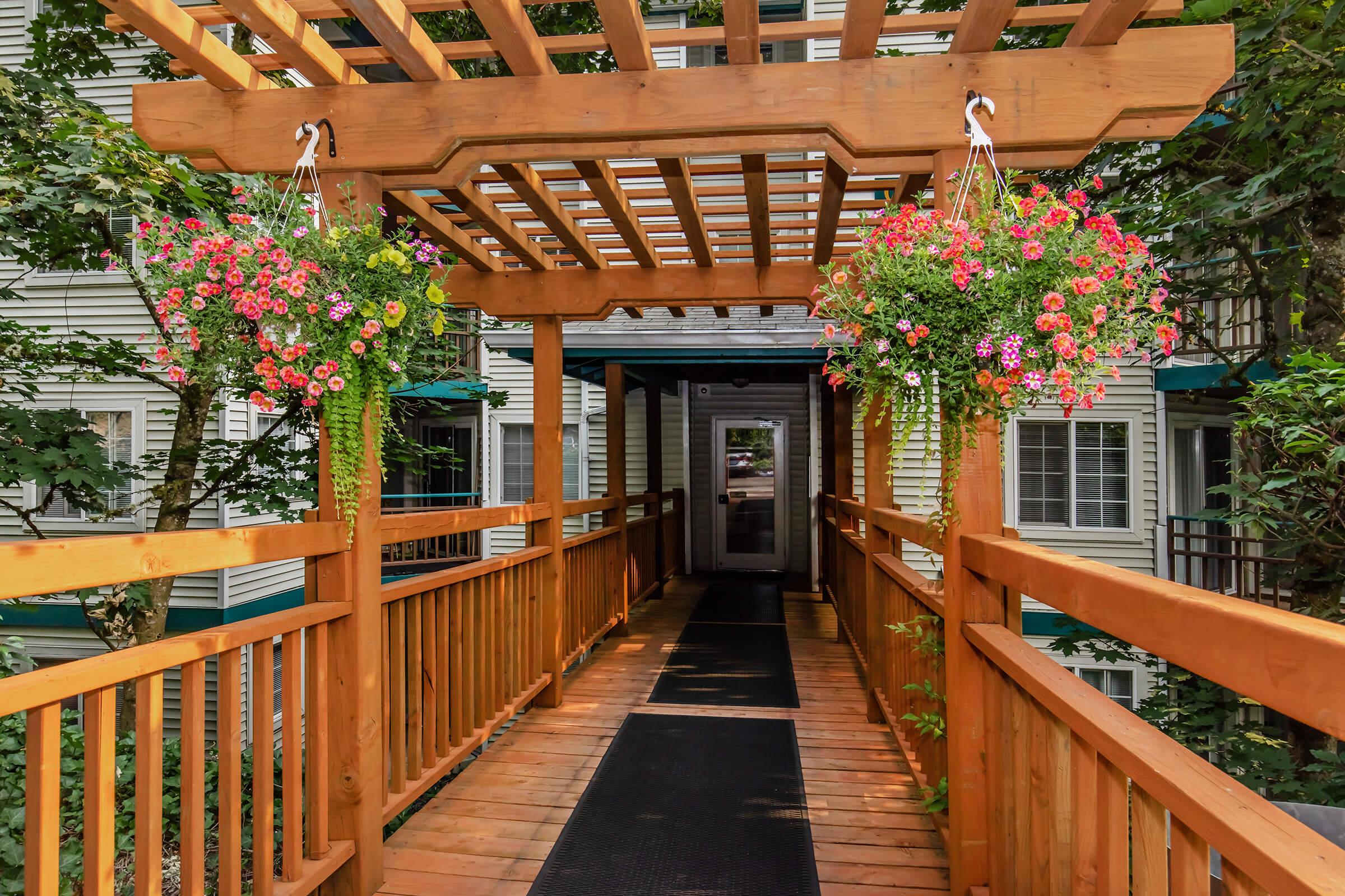 A wooden pergola with hanging flower baskets frames a pathway leading to a building entrance. The walkway is paved with black material and lined with greenery, creating a welcoming atmosphere. The surrounding architecture features light-colored siding and multiple windows.