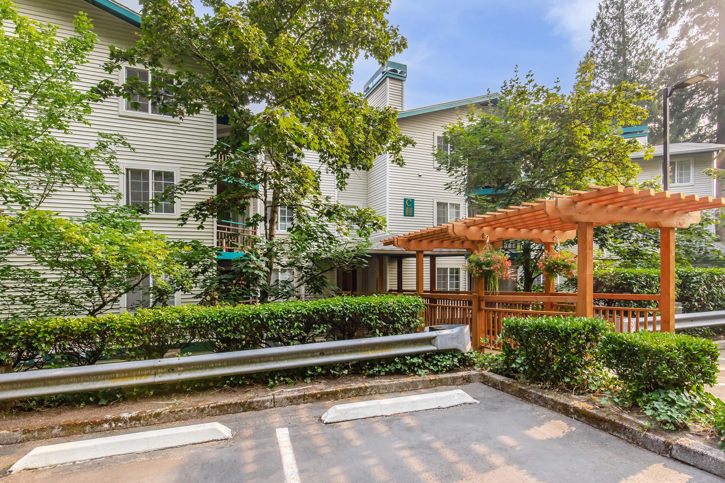 A view of a residential building surrounded by lush green trees and shrubs. In the foreground, there is a wooden pergola adorned with plants, and a parking area with empty spaces. The sky is clear with a few clouds, creating a tranquil and inviting atmosphere.