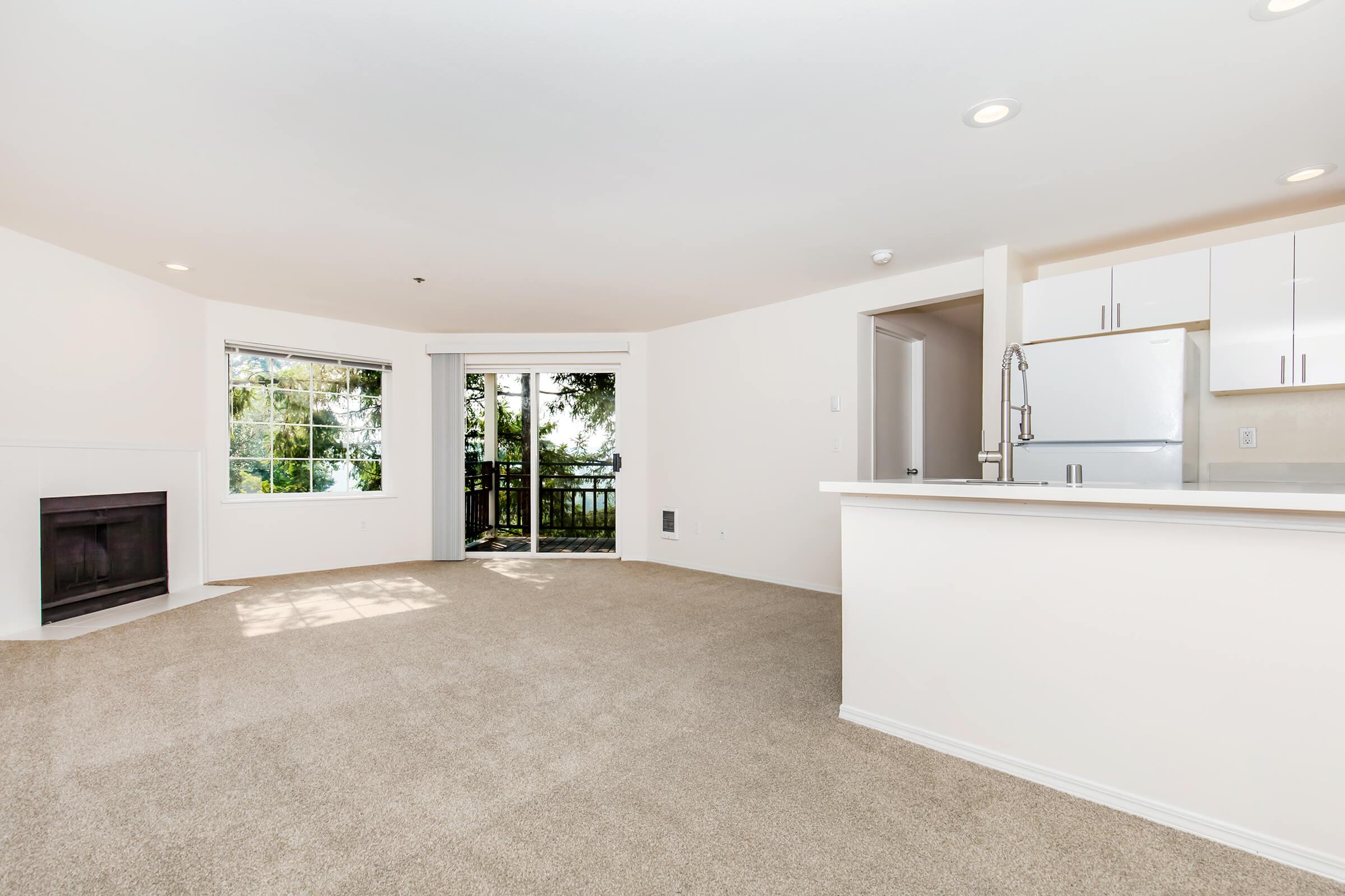 A spacious, bright living room with light-colored carpet and white walls. It features a large window with a view of greenery, a sliding glass door leading to a balcony, a modern kitchen with white cabinets, and a cozy corner fireplace. The area is well-lit with recessed ceiling lights.