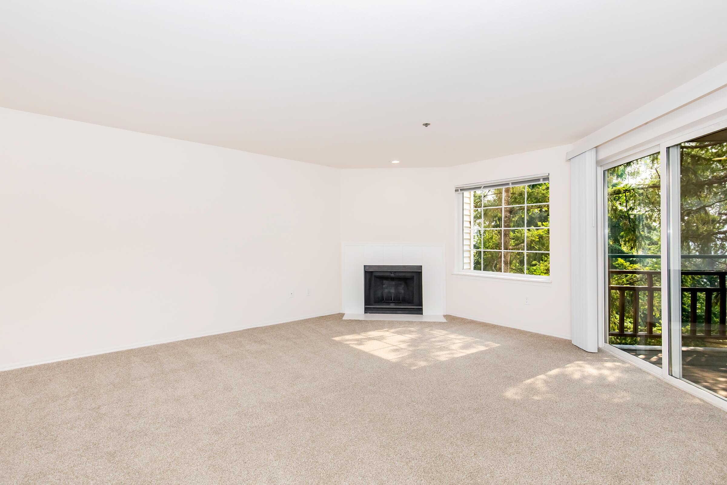 A bright and spacious living room featuring light beige carpet, a white fireplace, and large windows with views of the outdoors. The walls are painted in a soft neutral color, and there is a sliding glass door leading to a balcony area.