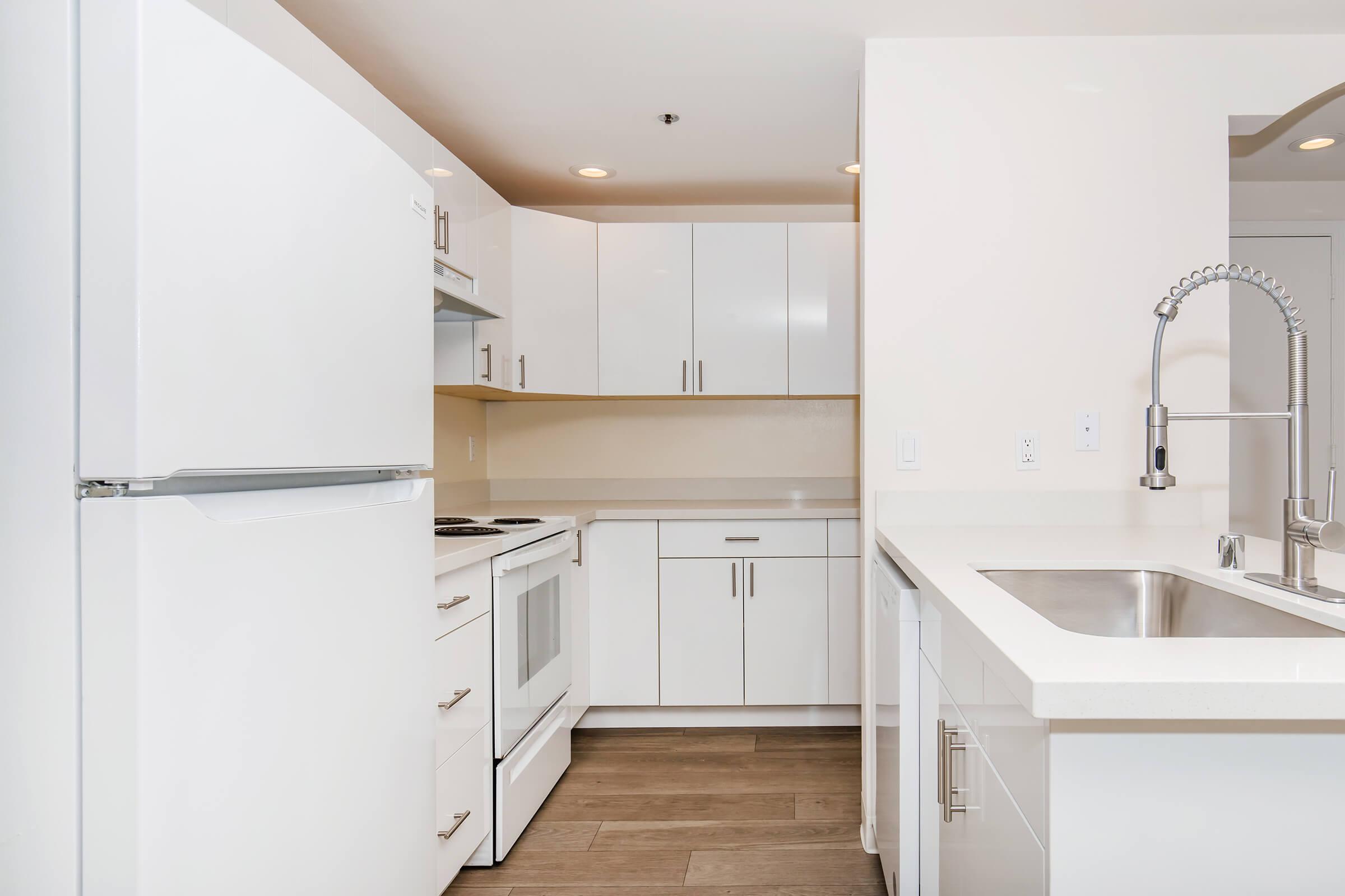 Modern kitchen featuring white cabinets and appliances. Includes a refrigerator, oven, and sink with a sleek faucet. The countertops are light-colored and the flooring is wood. The design emphasizes a clean, contemporary look with an open layout.