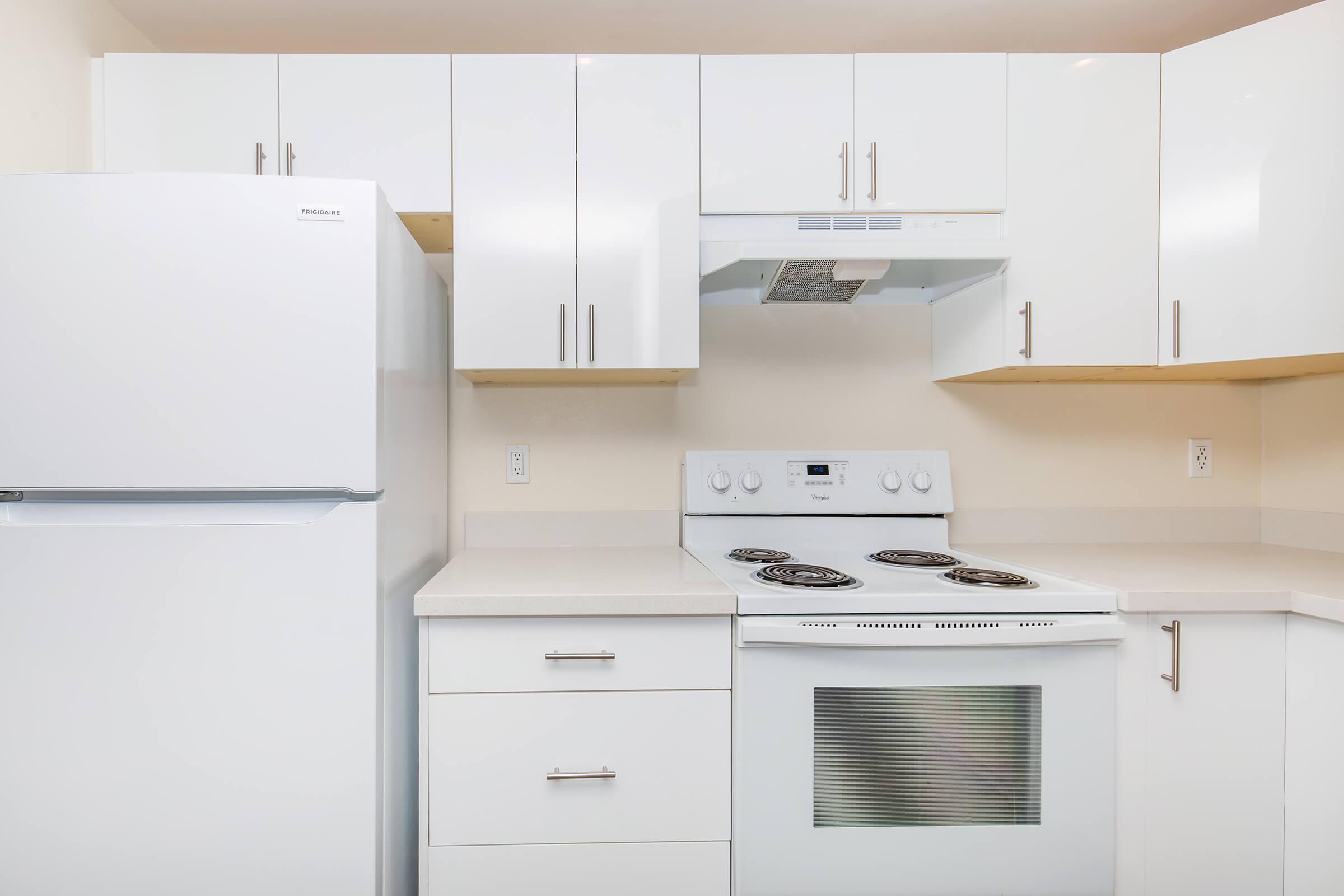 A modern kitchen featuring white cabinetry, a stainless steel fridge, a white stove with four burners, and a hood above the stove. The countertop is light-colored, and the overall design is sleek and contemporary, with minimal clutter and a bright, clean appearance.
