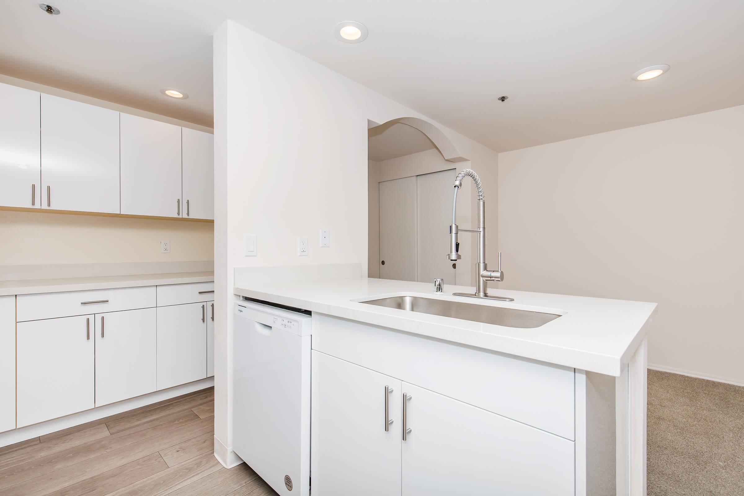 Modern kitchen featuring white cabinets and a sleek countertop with a sink and a pull-down faucet. A dishwasher is installed under the counter. The space has neutral-colored walls and laminate flooring, with ample lighting from recessed ceiling fixtures.
