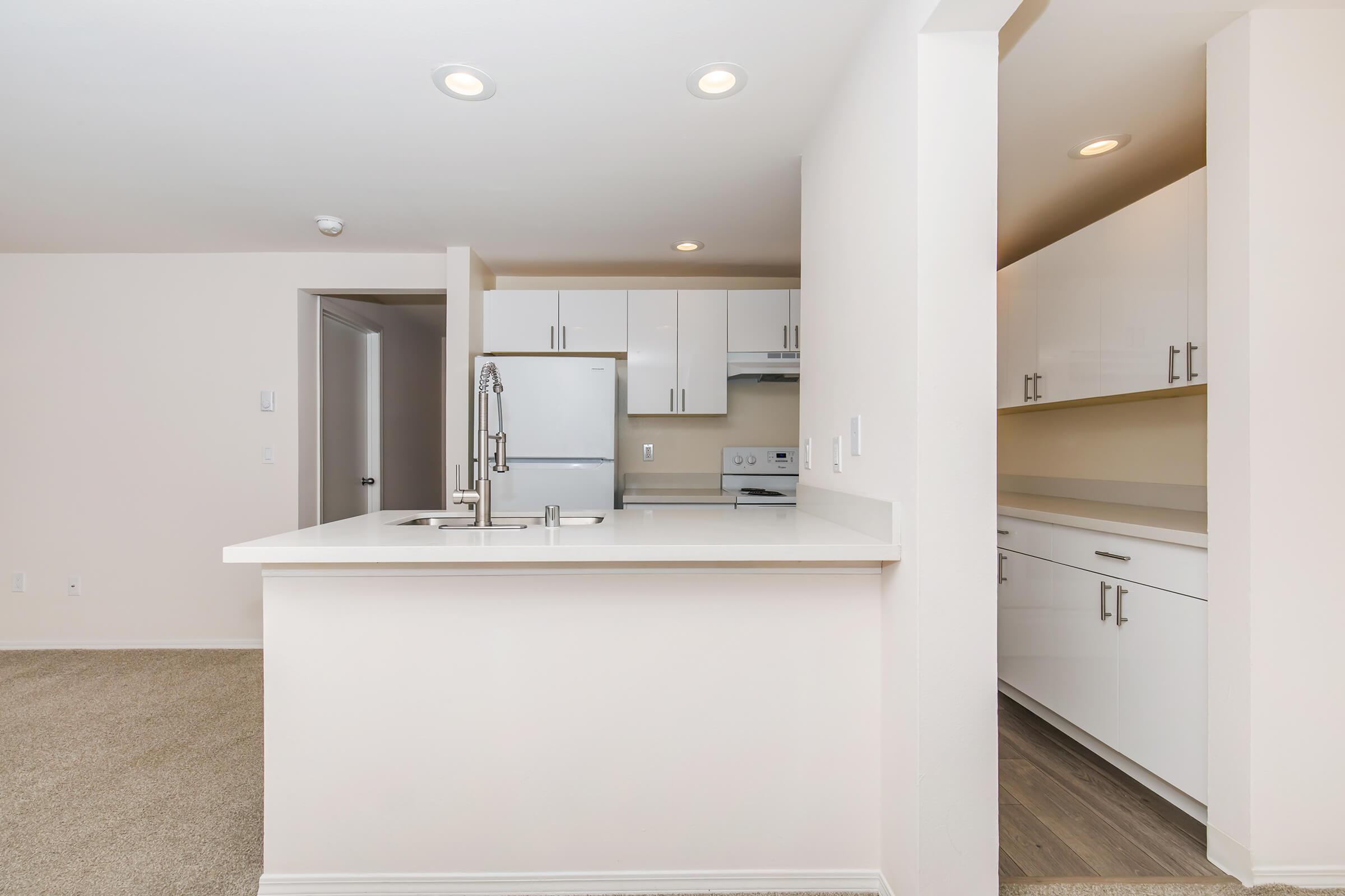 Modern kitchen with white cabinetry, stainless steel appliances including a refrigerator and stove, and a countertop with a sink. The space features neutral-colored walls and carpet flooring, creating a clean and inviting atmosphere. Bright lighting enhances the open layout.