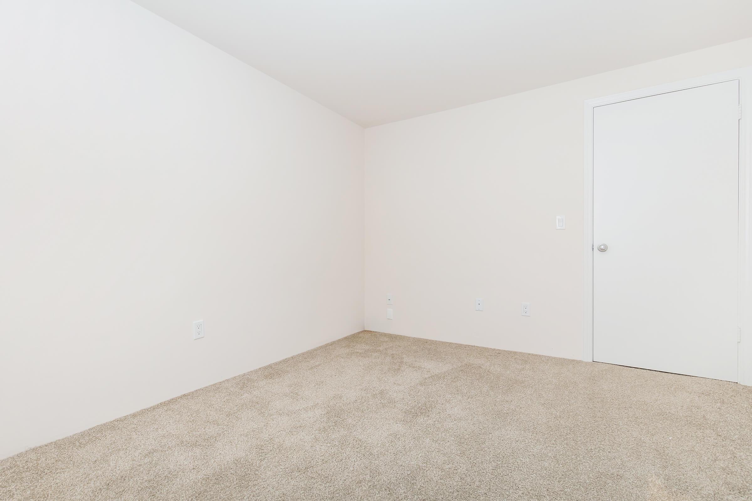 A clean, empty room with pale beige walls and light beige carpet. There is one white door on the right leading to another space, and no furniture or decorations present, creating a minimalistic and spacious atmosphere.