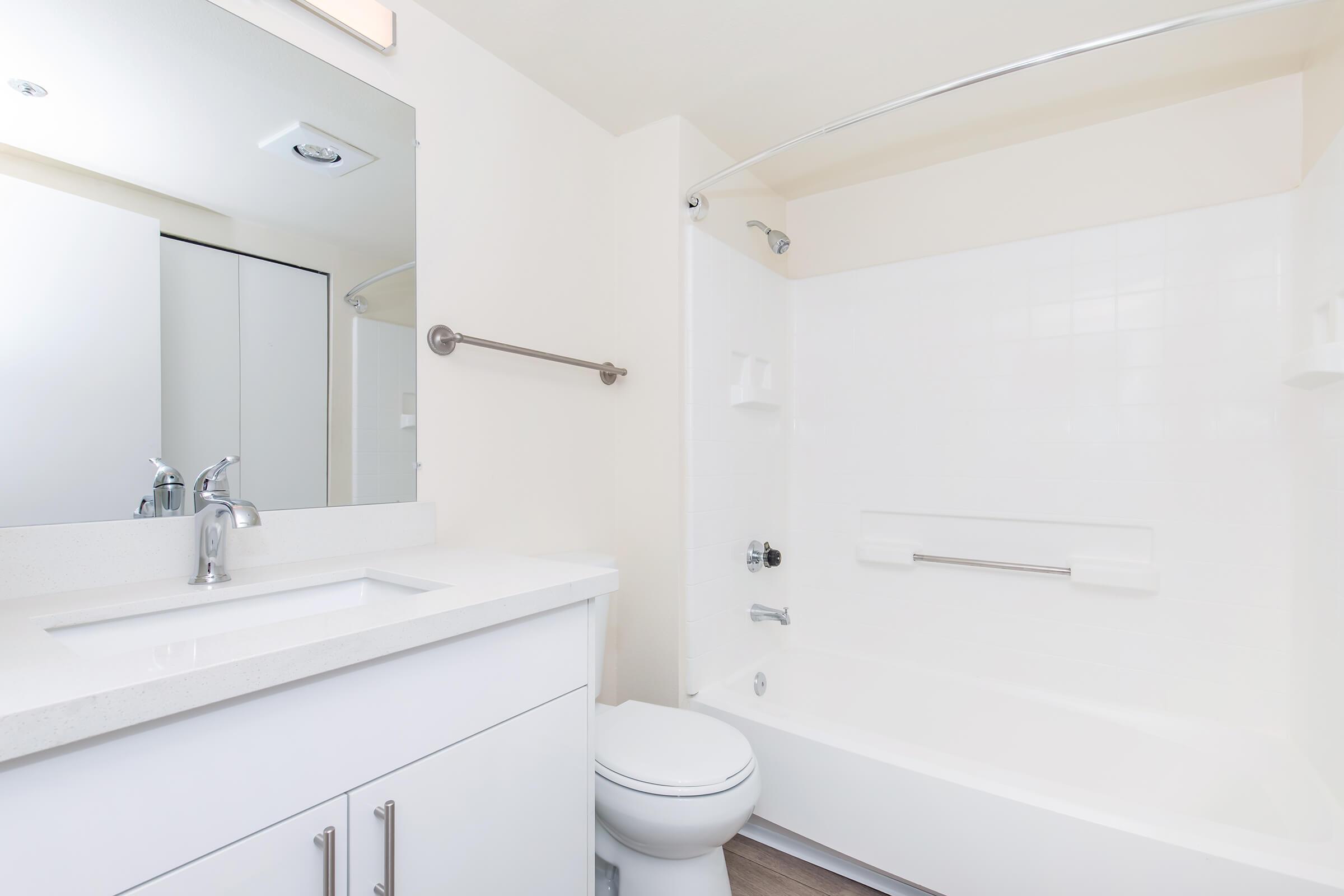 Modern bathroom featuring a white tub with an integral shower, a sleek white countertop with a sink, a large mirror above the sink, and a white cabinet. Light-colored walls and wood-like flooring create a bright and clean atmosphere.