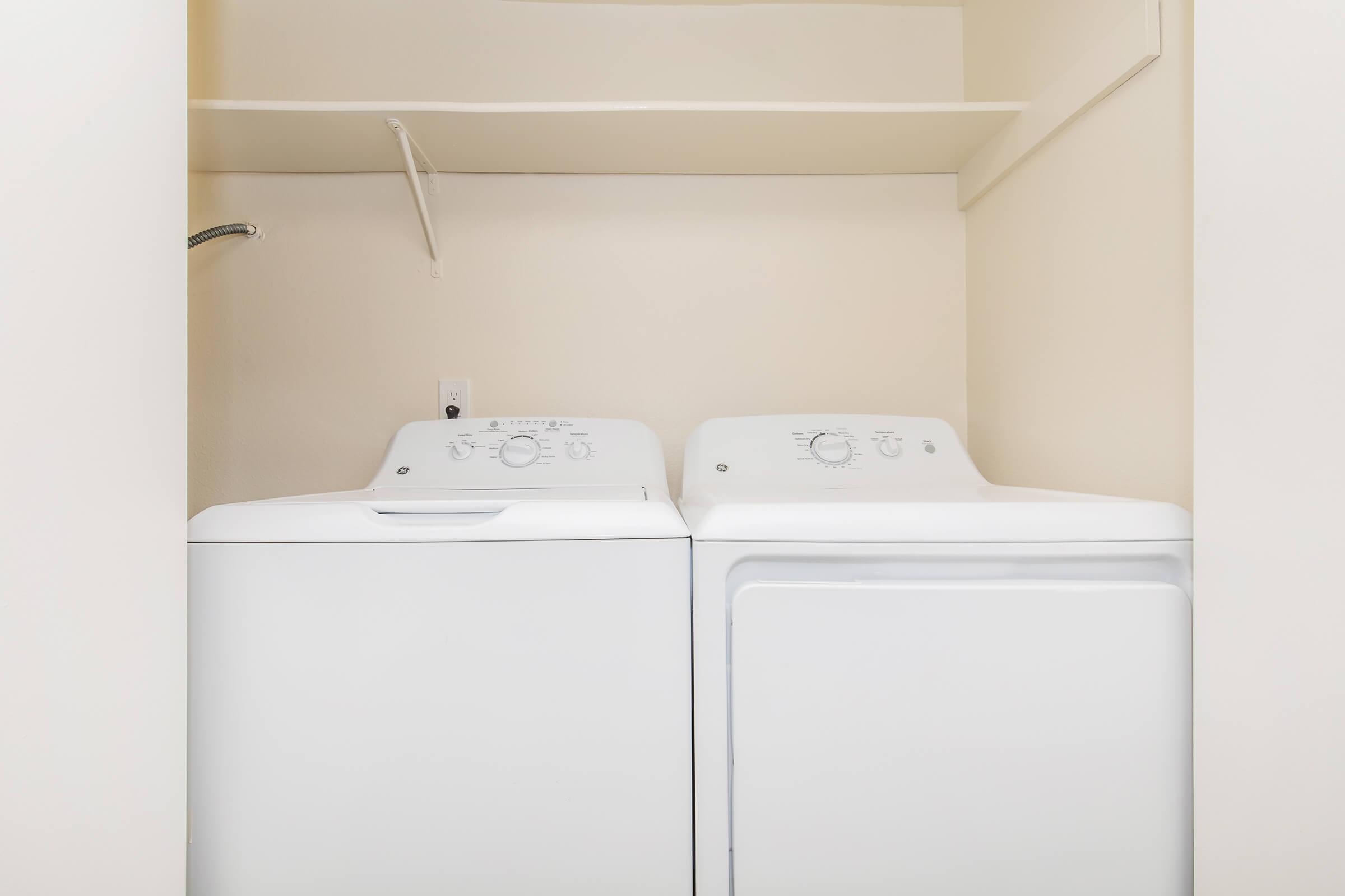 A clean, empty laundry room featuring a stacked washer and dryer unit against a light-colored wall. The space includes a small shelf above the appliances, providing storage options.
