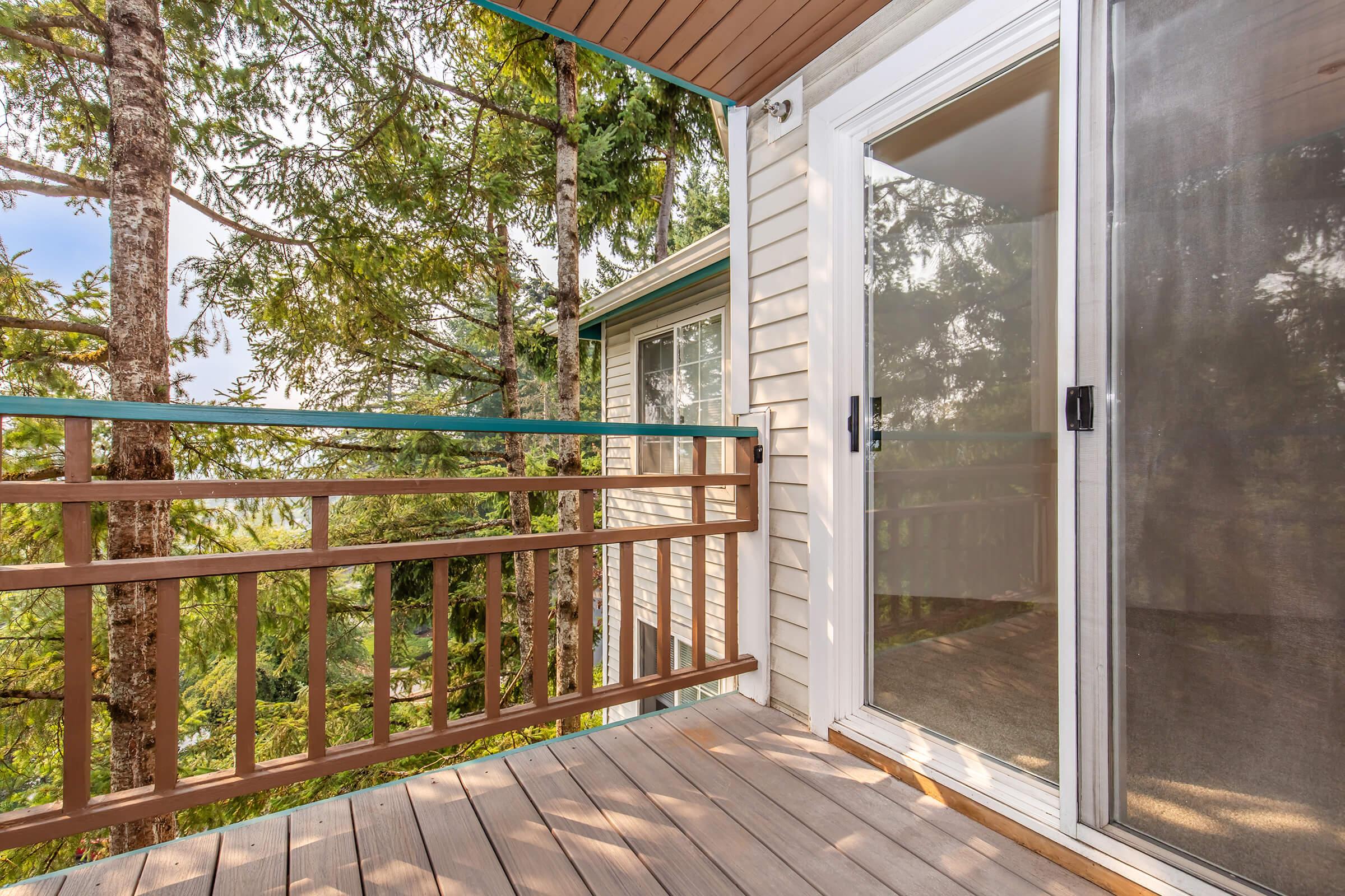 A wooden balcony with a railing, leading to a sliding glass door. The area is surrounded by tall trees and green foliage, creating a serene outdoor view. The floor is made of wooden planks, and the walls of the house are visible in the background, partially obscured by the trees.