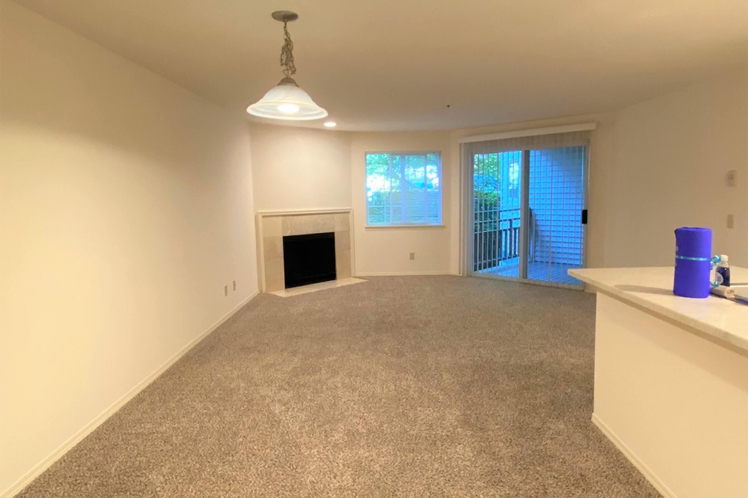 A spacious, unfurnished living room with light beige walls and gray carpet. There is a fireplace on the left, a large window providing natural light, and sliding glass doors leading to a patio area. A counter separates the kitchen from the living space, with a blue water bottle visible on the counter.