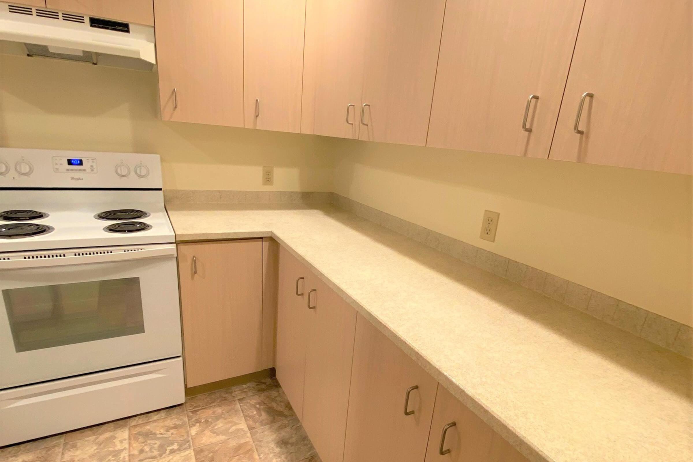 A clean, modern kitchen featuring light-colored cabinets, a white stove with four burners, and a beige countertop. The walls are painted a soft cream, and the floor has a patterned tile. There are no visible appliances or utensils on the countertop, creating a spacious and uncluttered appearance.