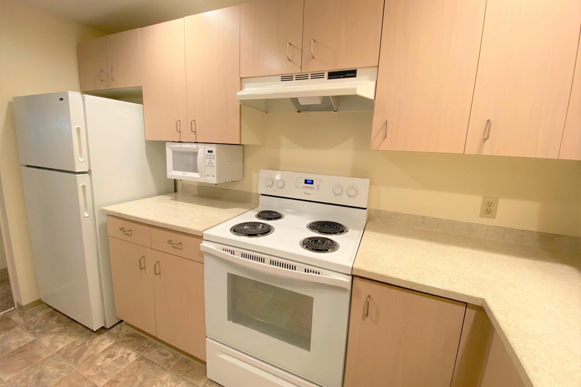 A modern kitchen featuring light wood cabinetry, a white refrigerator, a microwave, and a white stove with four burners. The countertop is a neutral color, and the floor has tiles in a subtle pattern. The kitchen is well-lit, providing a clean and functional cooking space.