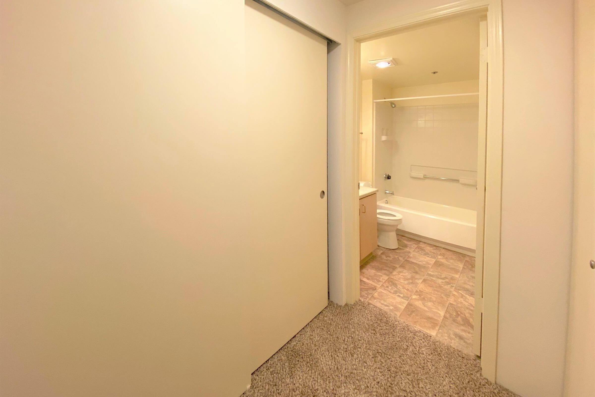 A view of a bathroom from a hallway, featuring light-colored walls and carpet. The bathroom includes a bathtub, toilet, and sink. A closed sliding door is visible on the left side, and there is natural light illuminating the space.