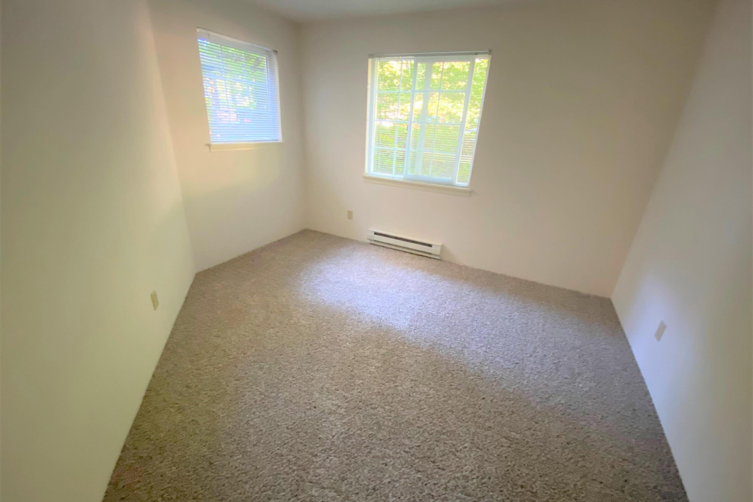 A vacant room with light beige carpet and white walls. There is a window with blinds allowing natural light to enter. The room is empty, creating a spacious and airy feel. A baseboard heater is visible beneath the window.