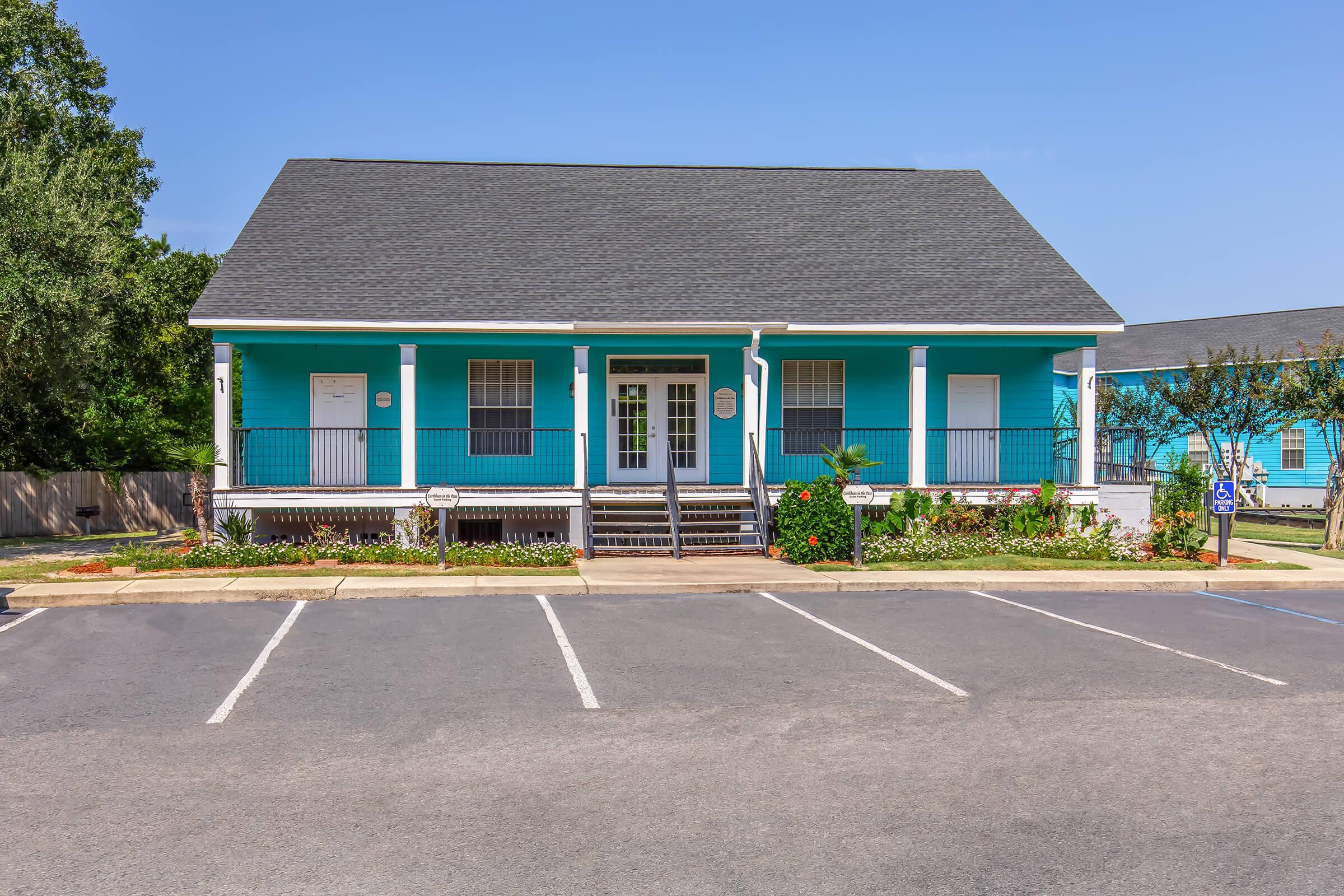 Single-story building with a bright turquoise exterior and a gray shingle roof. It has white trim and a front porch with steps leading up to the entrance. Surrounding the building are landscaped flower beds and a parking lot with empty spaces. The sky is clear and blue.