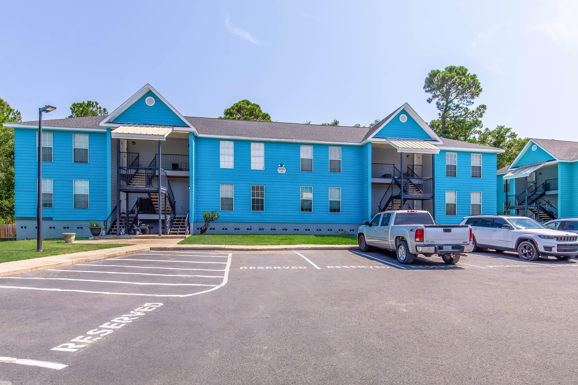 A bright blue apartment building with two levels, featuring staircases on either side. The parking lot in front shows several parked vehicles, including a white truck and an SUV. Green trees line the background under a clear blue sky. Reserved parking spaces are marked.