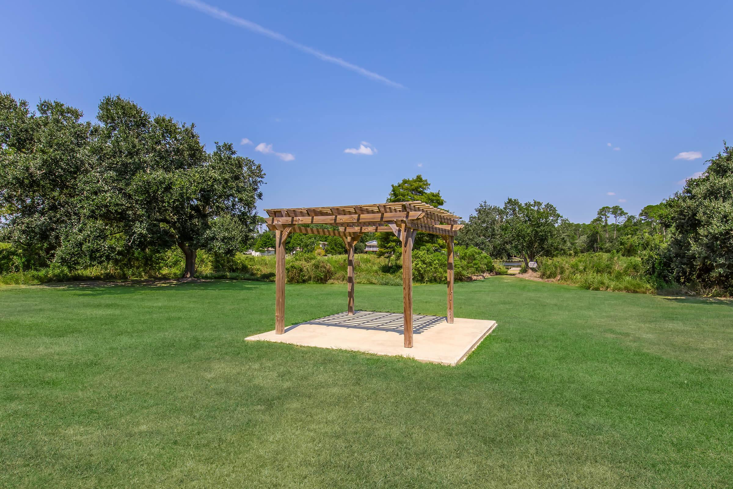 A wooden gazebo structure with a slatted roof is set on a grassy area, surrounded by trees under a clear blue sky. The gazebo has a concrete base and is situated in a well-maintained outdoor space, ideal for relaxation or gatherings.