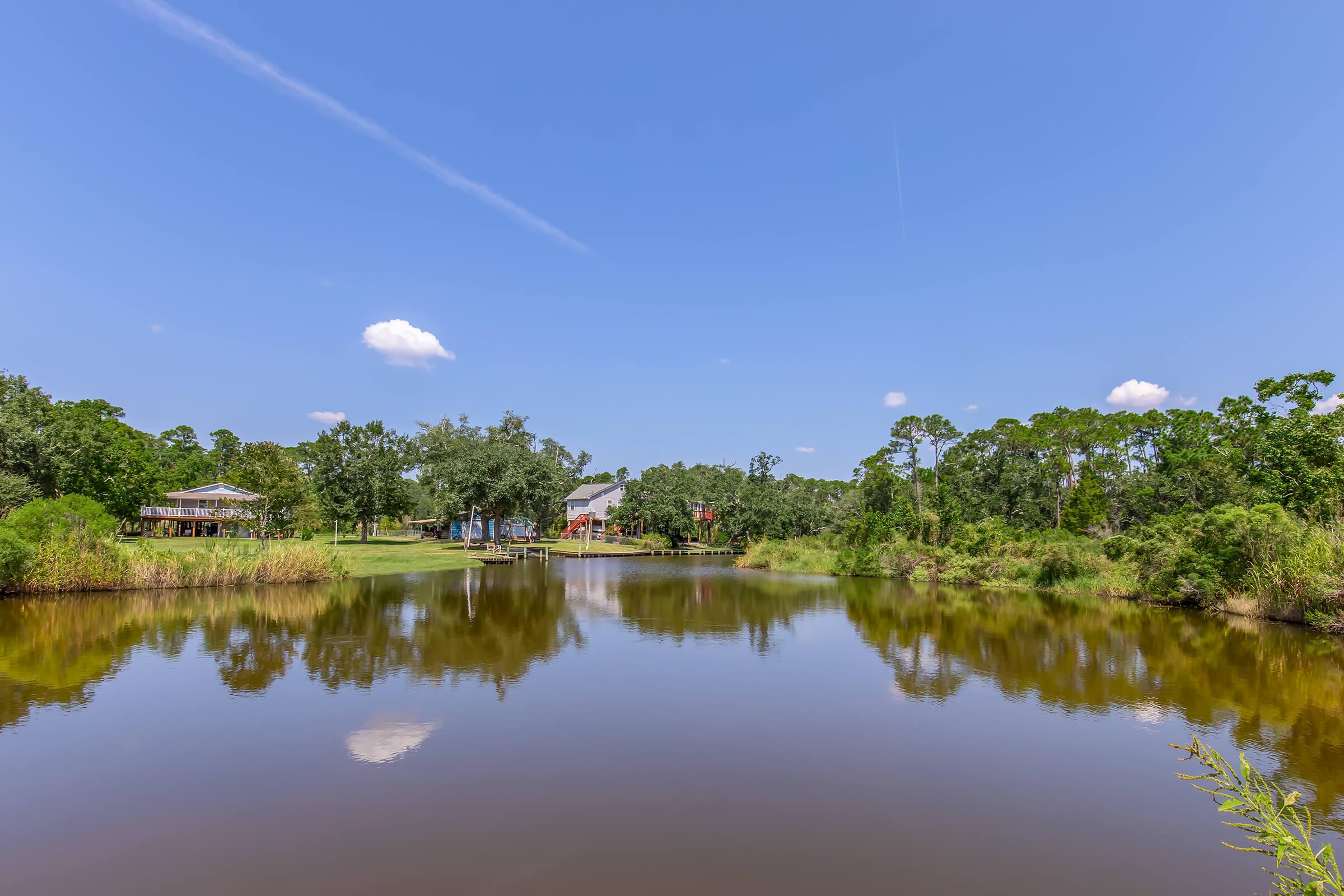 A serene landscape featuring a calm pond reflecting the blue sky and fluffy white clouds. Lush greenery surrounds the water, with trees and a grassy area visible. In the background, there are structures and shelters, enhancing the peaceful atmosphere of the natural setting.