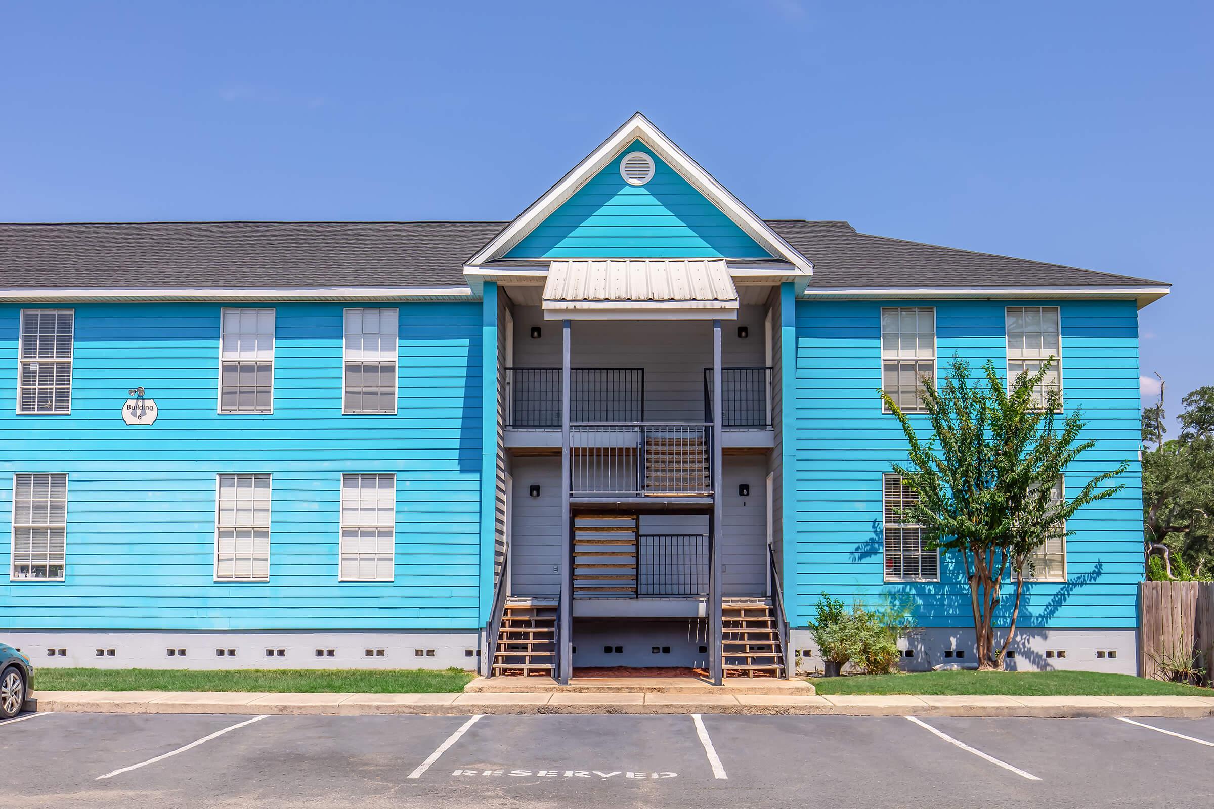 A bright blue apartment building with white trim, featuring two levels. The front has a central entrance with stairs leading up to a covered porch. There are large windows and a small green area with shrubs. Parking spaces are marked in front, with one labeled "Reserved." The sky is clear and blue.