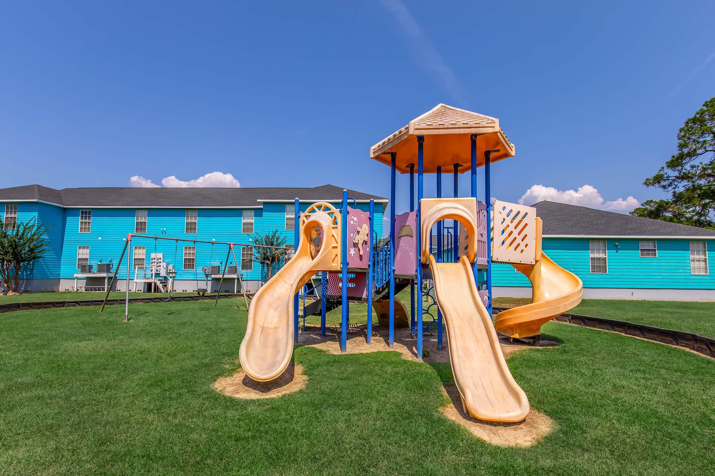 A colorful playground with a slide, climbing structure, and swings, set on vibrant green grass. Behind it, there's a bright blue building under a clear sky with a few clouds. The scene conveys a cheerful, inviting atmosphere for children to play.