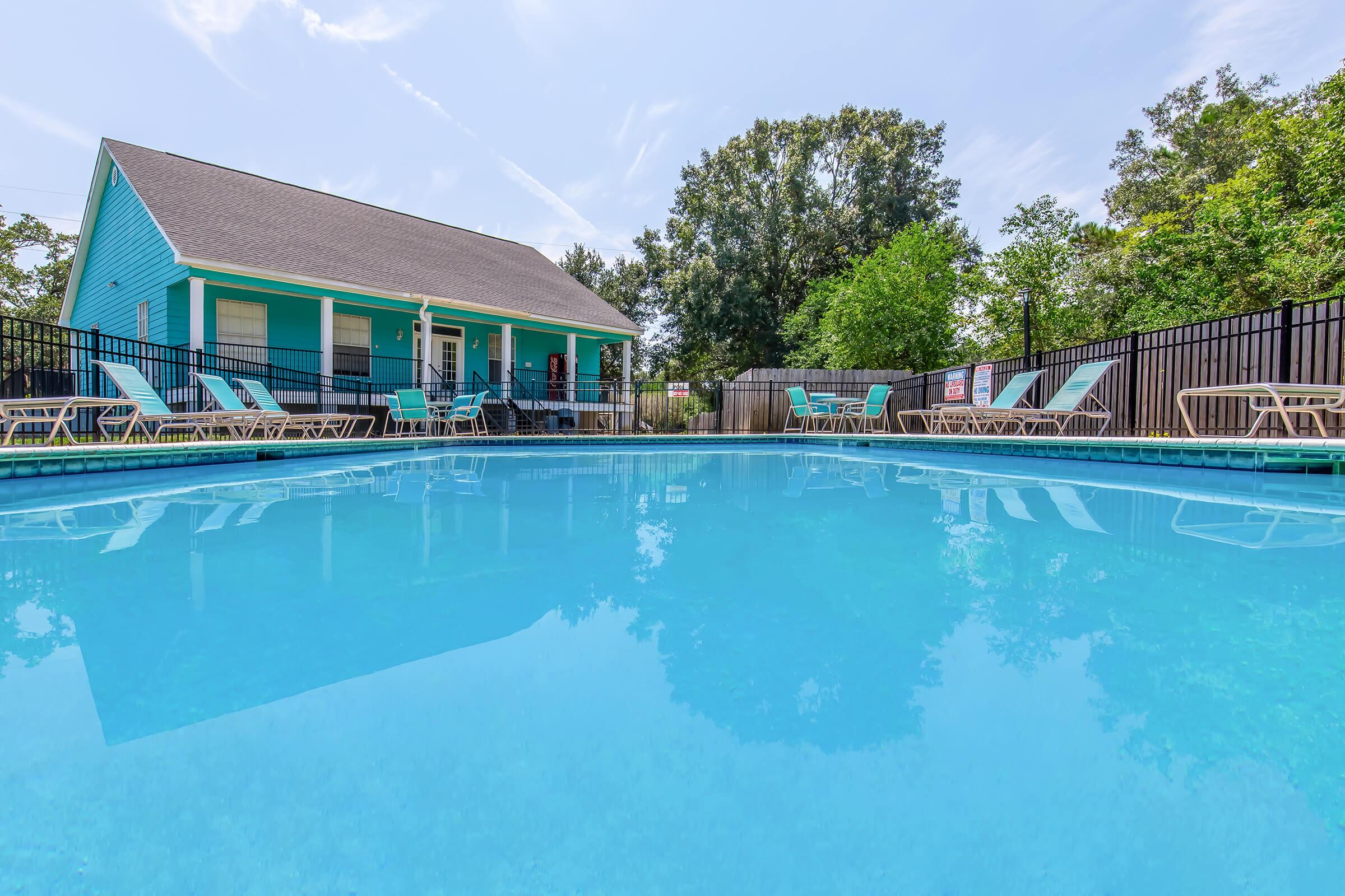 A sparkling blue swimming pool surrounded by lounge chairs, with a light blue house in the background. Lush green trees and blue skies create a relaxing atmosphere. Fences frame the pool area, enhancing the privacy of the space. Ideal for leisure and outdoor gatherings.