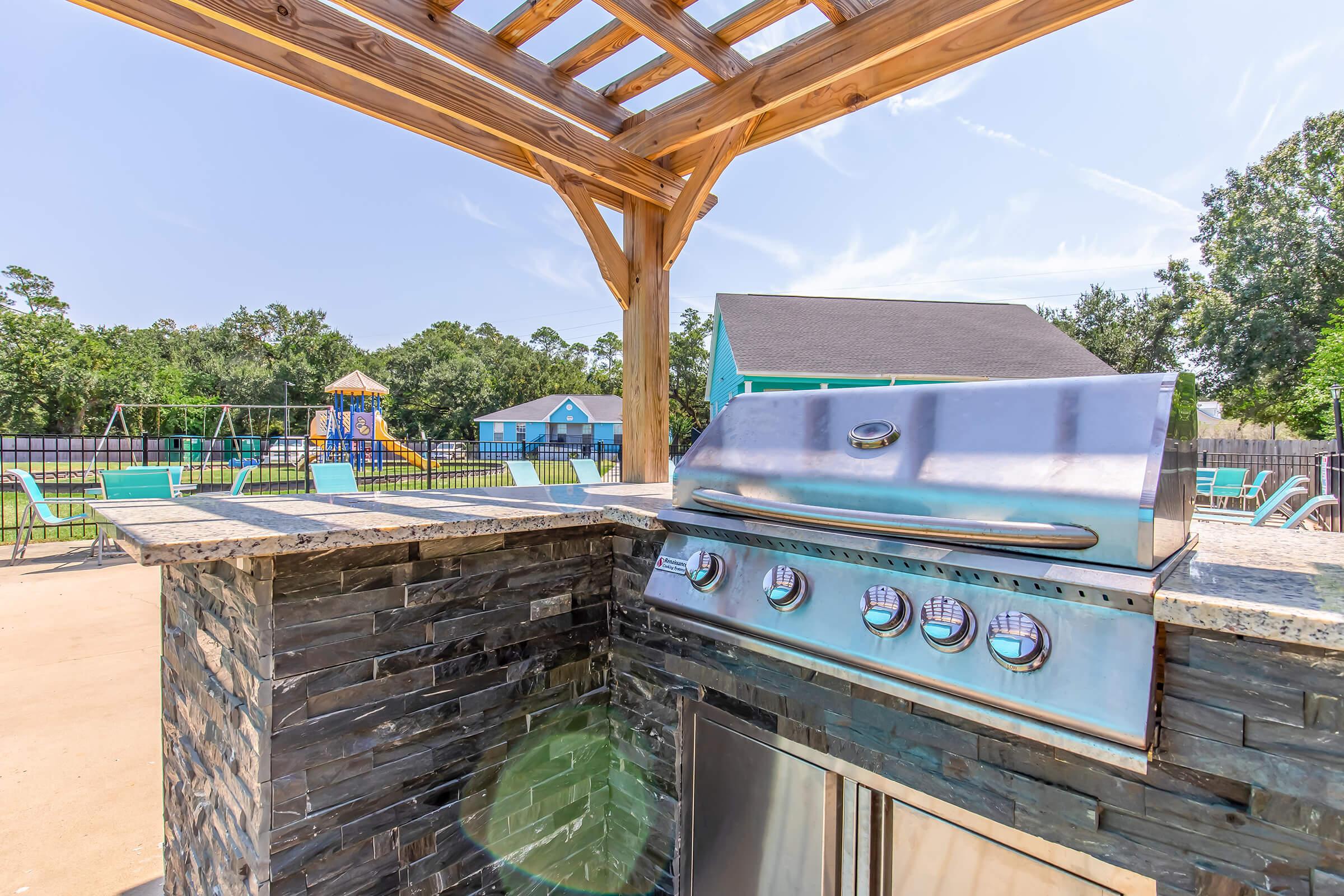 A modern outdoor grill under a wooden gazebo, set beside a swimming pool area. In the background, there are playground equipment and a colorful building, surrounded by trees. The scene is sunny and inviting, ideal for outdoor gatherings and barbecues.