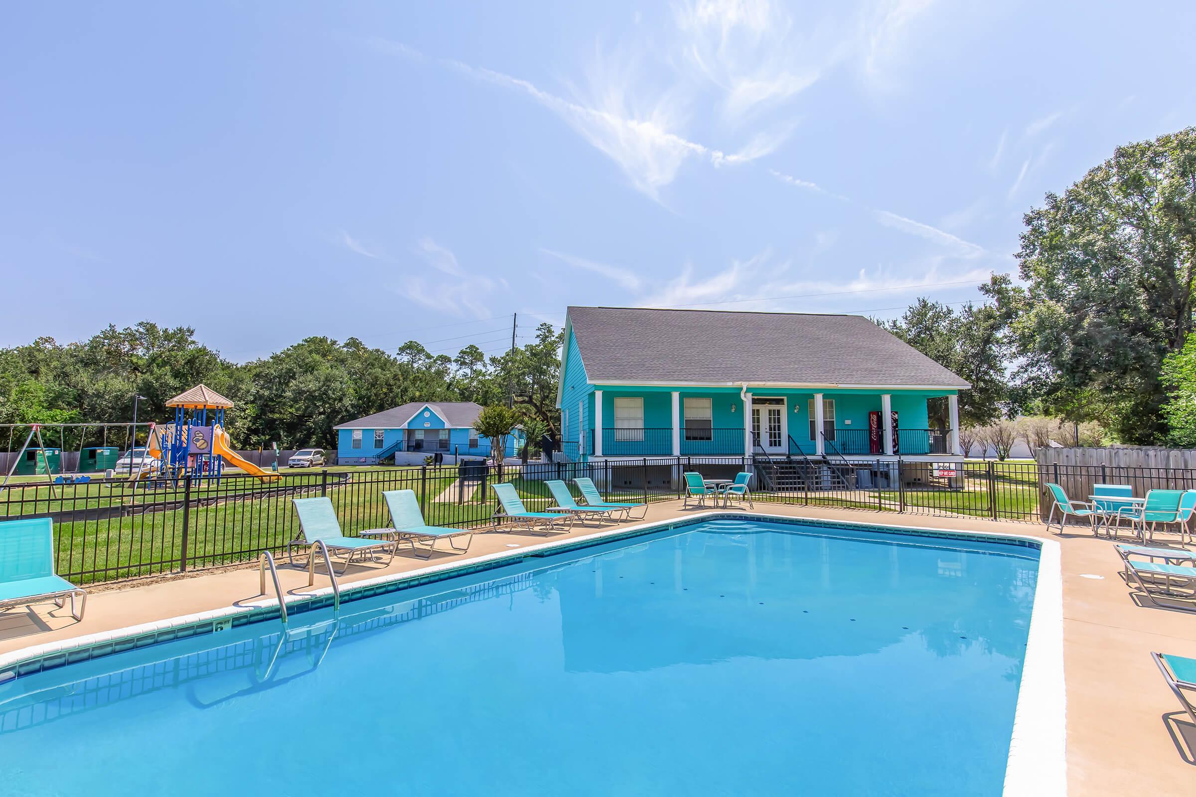 A vibrant outdoor scene featuring a clear blue pool surrounded by lounge chairs. In the background, a charming blue house and a playground are visible, set against a backdrop of greenery and a bright sky with wispy clouds.