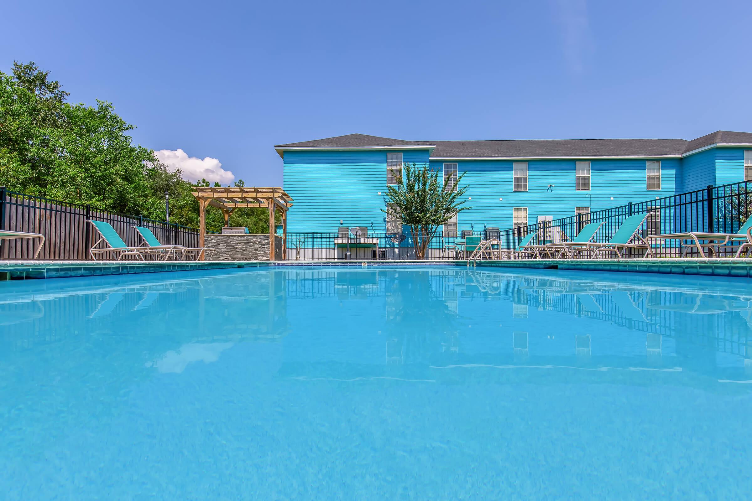 A clear blue swimming pool with lounge chairs around it, set against a vibrant blue building. The scene is bright and sunny, with a few trees and a pergola in the background, creating a relaxing outdoor atmosphere.