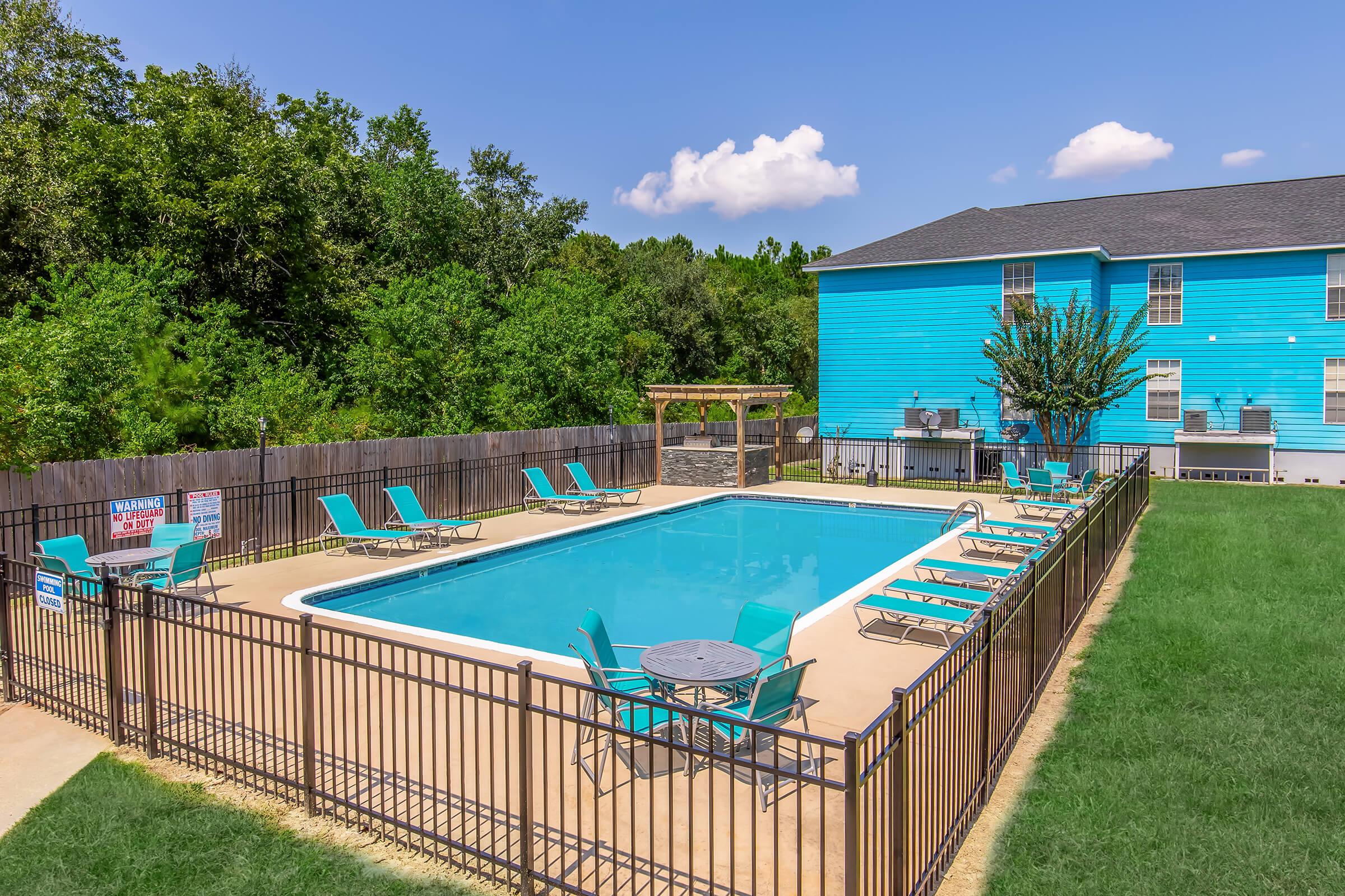 A clear blue swimming pool surrounded by a black metal fence. Lounge chairs are positioned around the pool on a sandy area, with a small table in the center. The background features a vibrant blue building and lush greenery under a bright sky with a few clouds.
