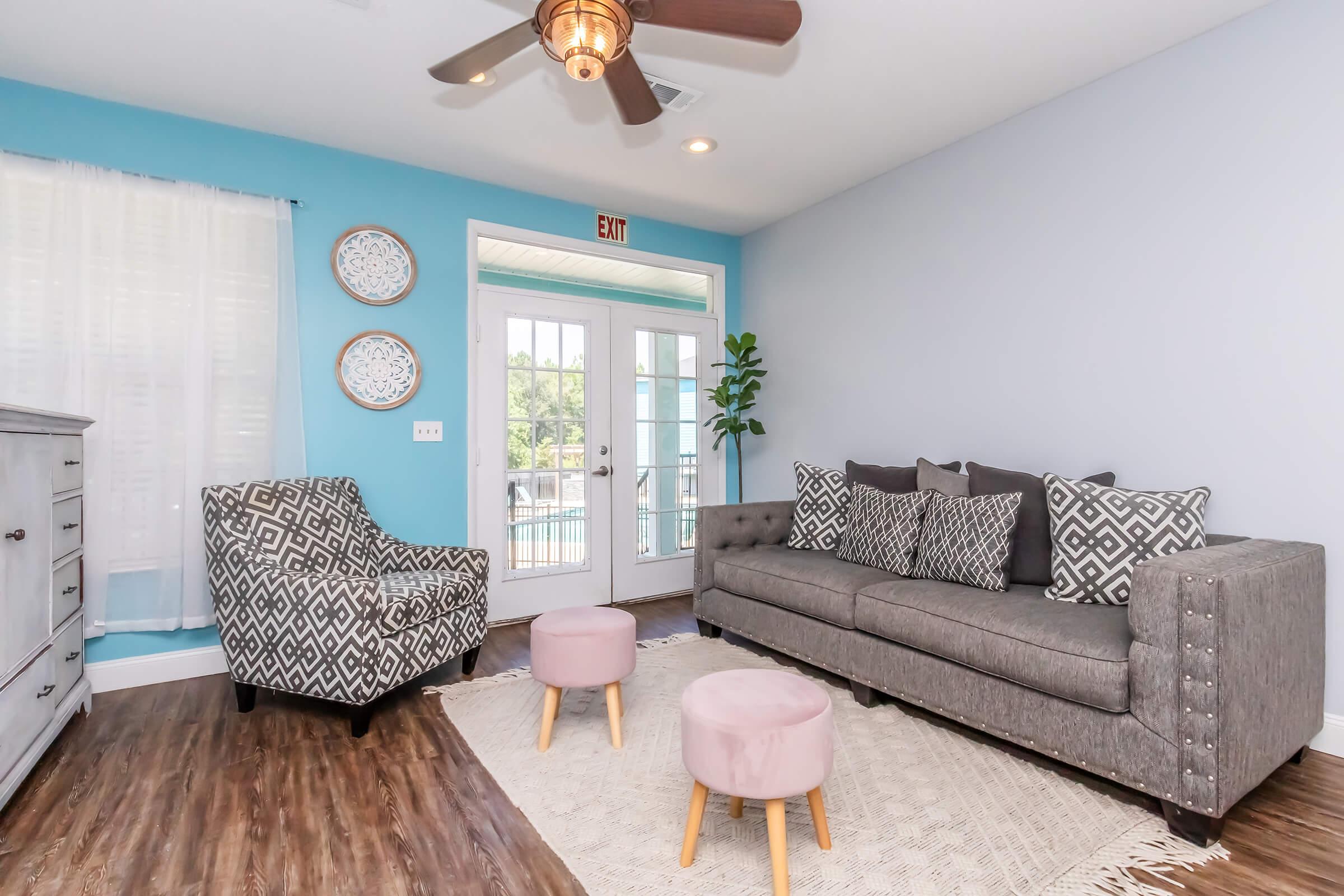 A modern living room featuring a gray sofa with decorative pillows, a patterned armchair, and two small pink stools. Natural light enters through sliding glass doors, illuminating the space. The walls are painted light blue, and there is a neutral area rug on the wood flooring, along with wall decor.