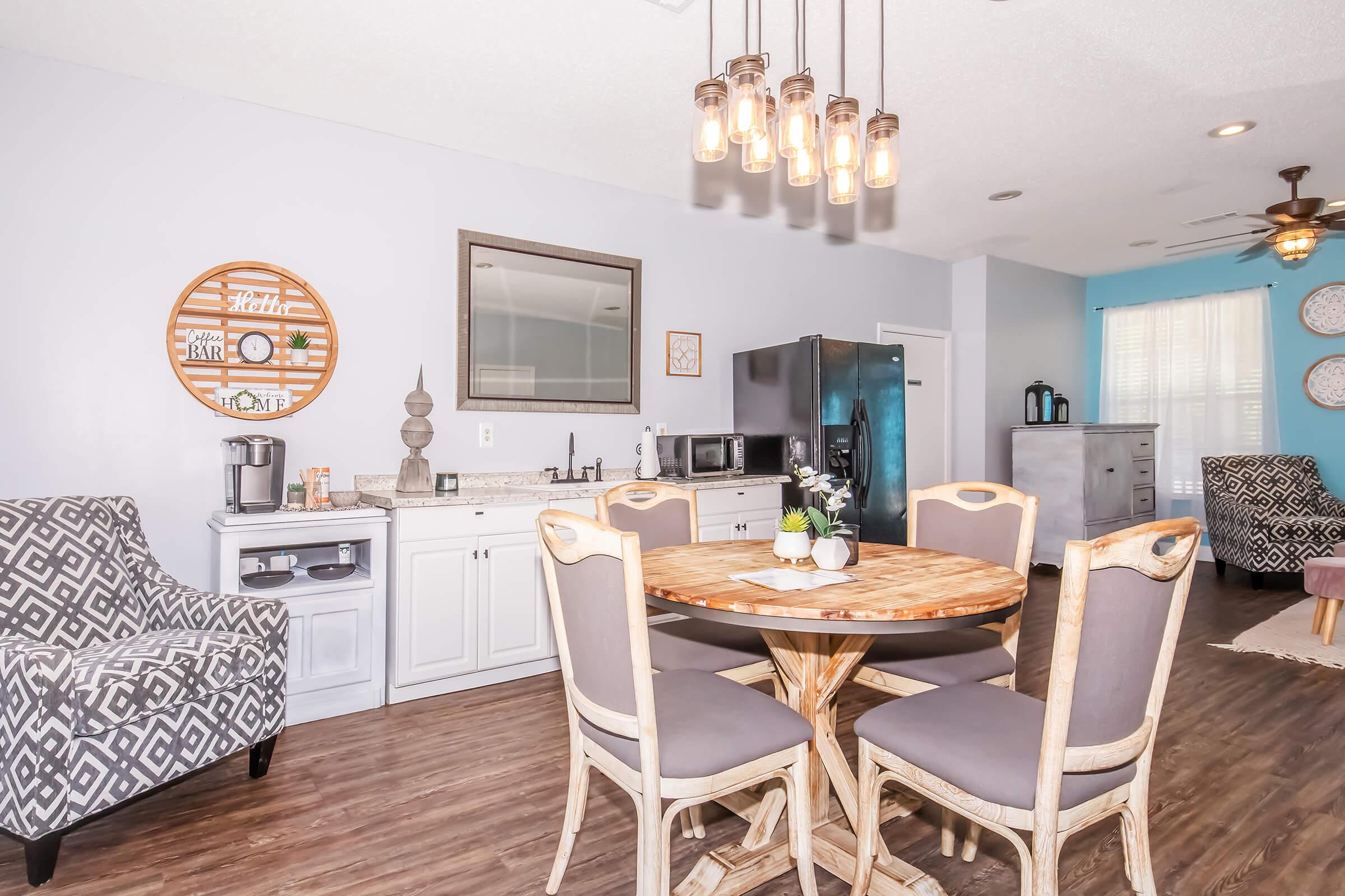 A modern kitchen and dining area featuring a round wooden table with six upholstered chairs, a light fixture with hanging bulbs above, a gray refrigerator, a countertop with a coffee maker and microwave, and a decorative wall mirror. The walls are painted light blue, and there are patterned chairs in the background.