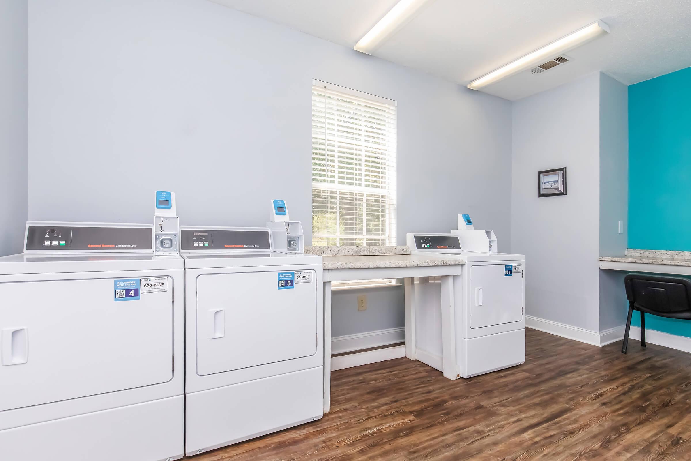 A clean and modern laundry room featuring two white washing machines and two white dryers. The room is well-lit with natural light coming through a window, and has light gray walls. A countertop provides space for folding clothes, and a black chair is visible in the corner.