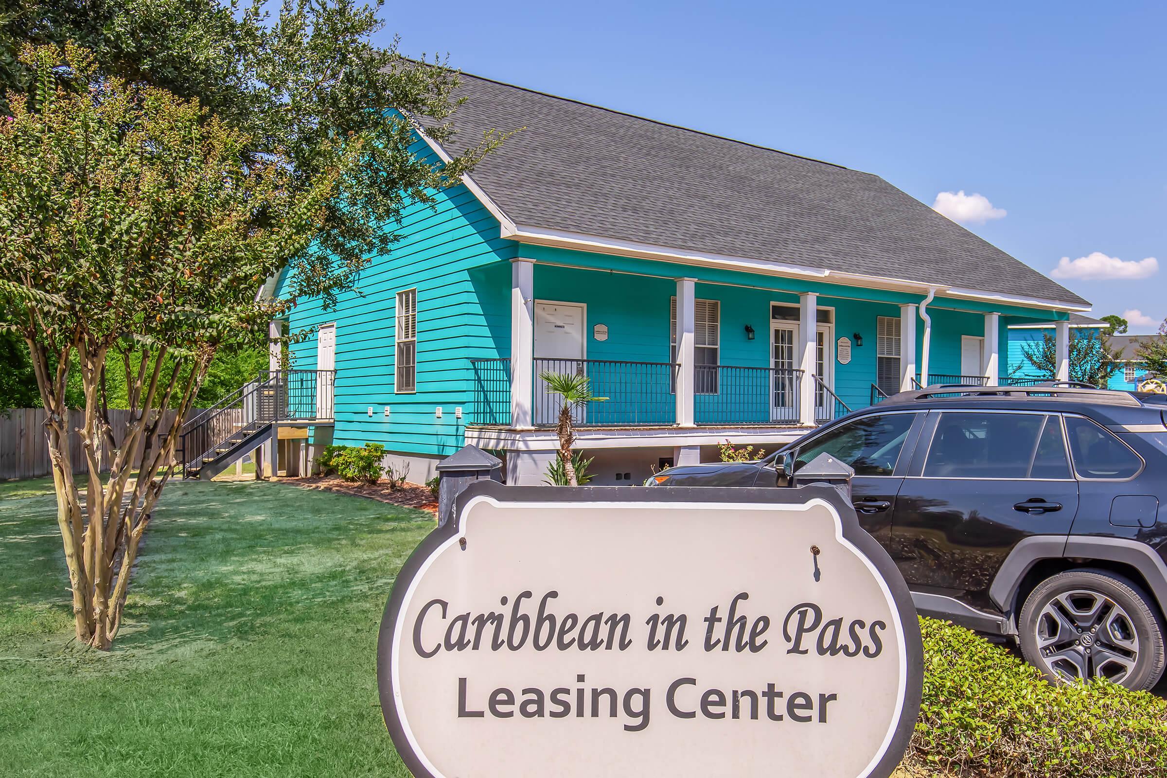 A bright turquoise building with a black roof, serving as the Leasing Center for "Caribbean in the Pass." The front features a sign with the name, surrounded by greenery and a parked car in the foreground. The sky is clear and blue, adding to the cheerful atmosphere of the location.