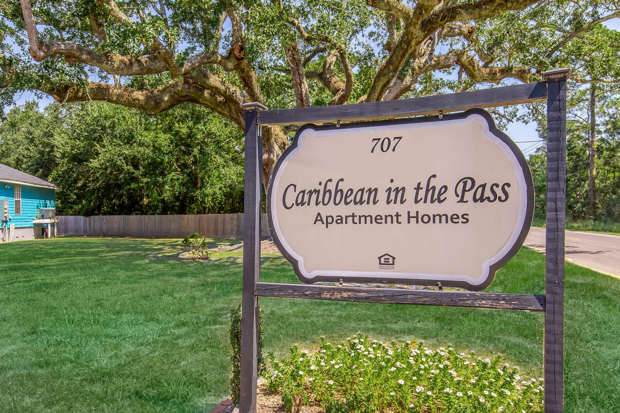 Sign for "Caribbean in the Pass Apartment Homes" located at 707. The sign is framed and surrounded by green grass and trees, with a clear blue sky above. The area appears well-maintained and inviting.