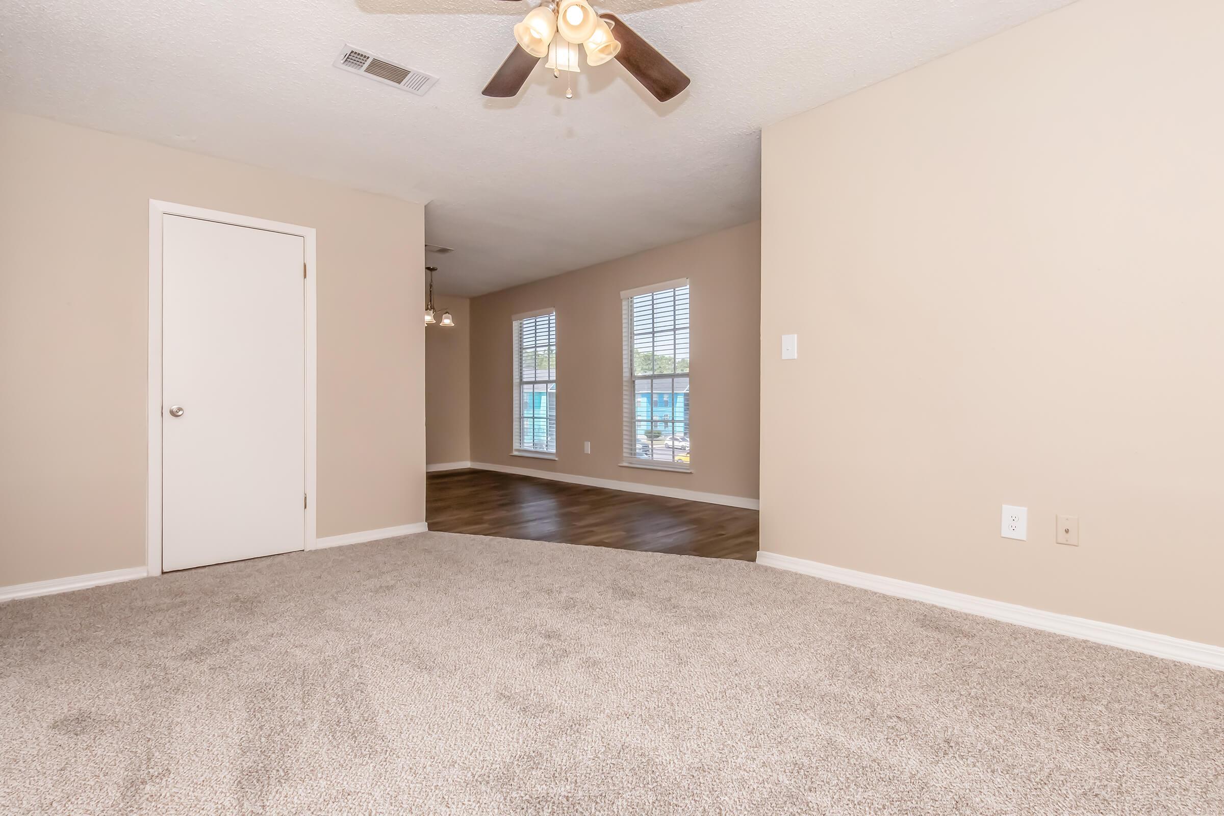 Spacious living area with beige walls and new carpet, featuring a ceiling fan. A doorway leads to another room with wooden flooring and large windows allowing natural light. The overall ambiance is inviting and bright, making it a perfect space for relaxation or gatherings.