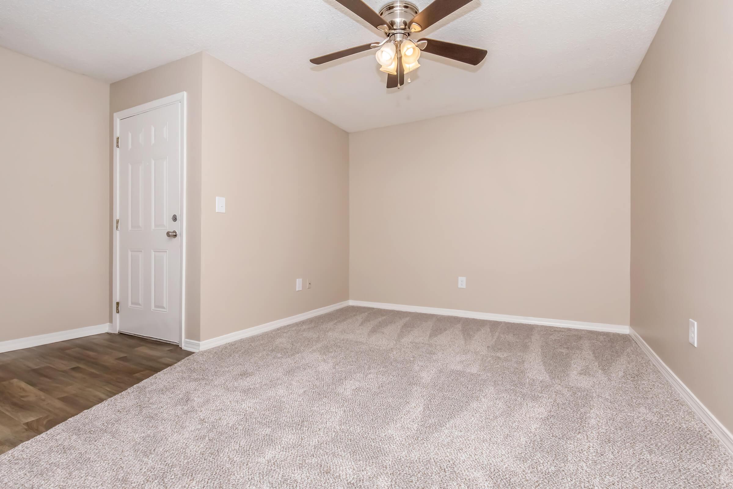 A vacant room featuring beige walls and carpeted flooring. A ceiling fan hangs in the center, and there is a white door leading to the outside. The room is well-lit with natural light and shows no furniture, emphasizing its spaciousness.