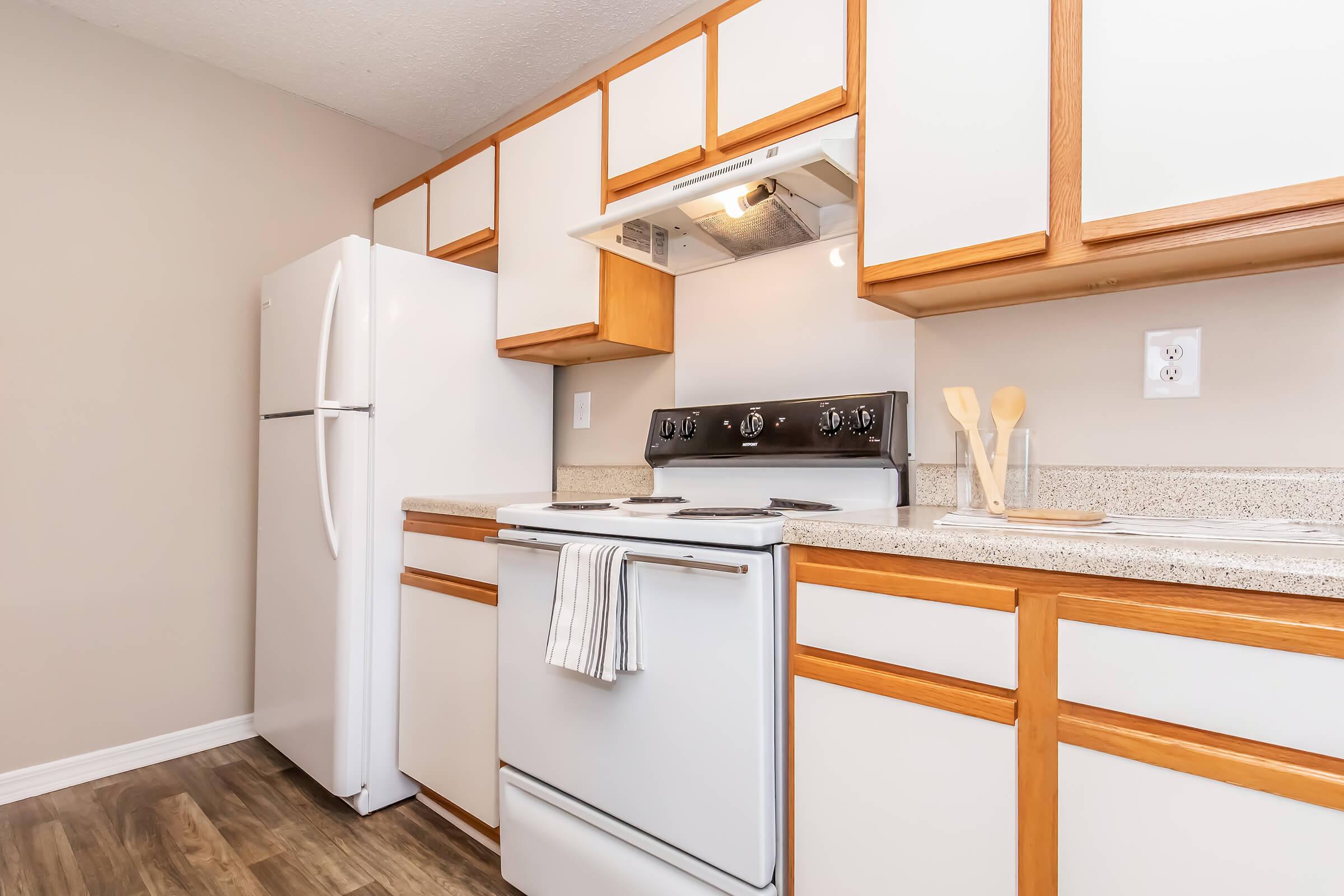 A modern kitchen featuring wooden cabinets, a white refrigerator, a stove with an overhead microwave, and a countertop. Kitchen utensils are visible, with a neutral wall color and laminate flooring, creating a clean and inviting space.